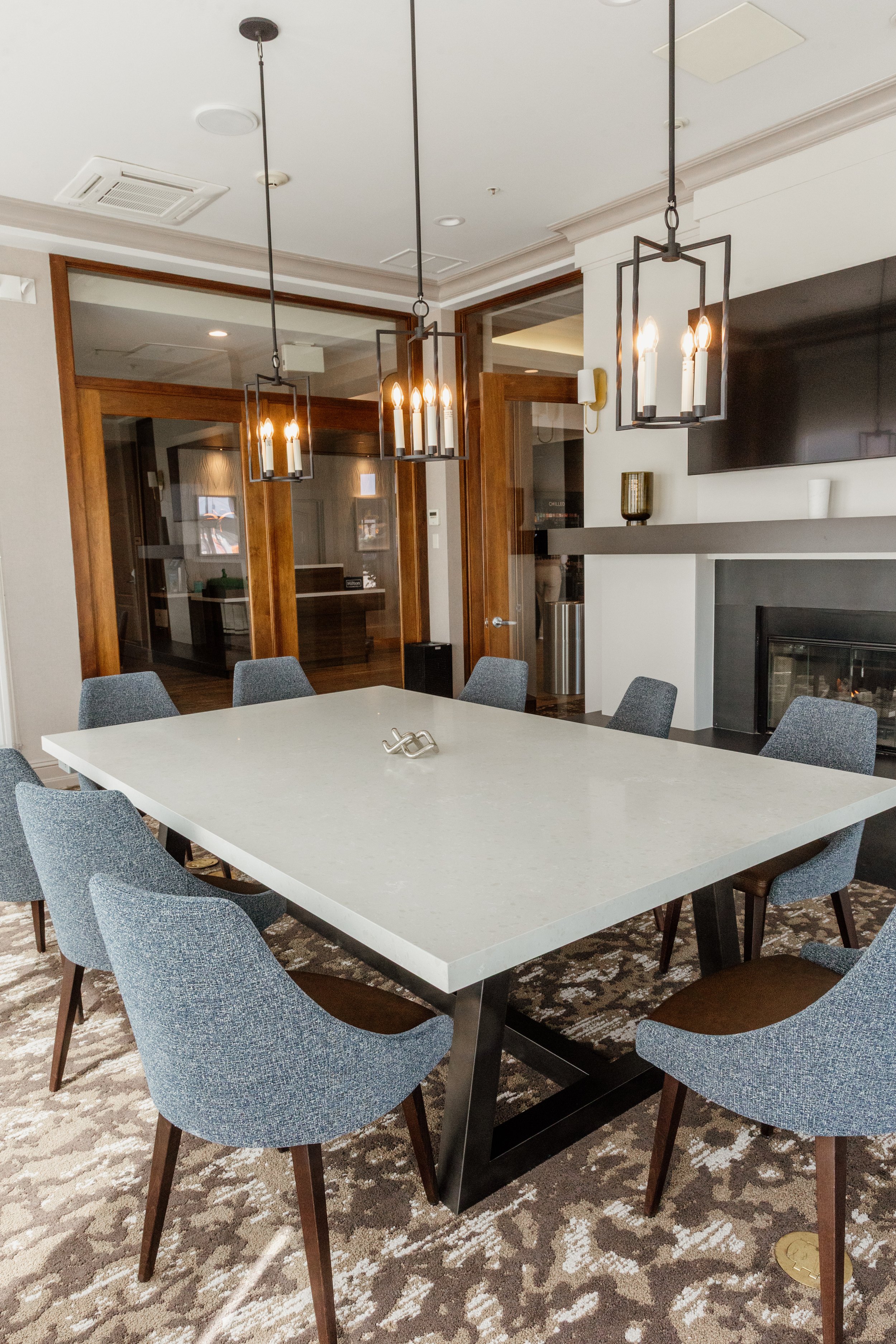 Modern dining room with a white rectangular table surrounded by eight blue upholstered chairs, two black metal pendant lights hanging above the table, a fireplace, and a flat-screen TV on the wall.