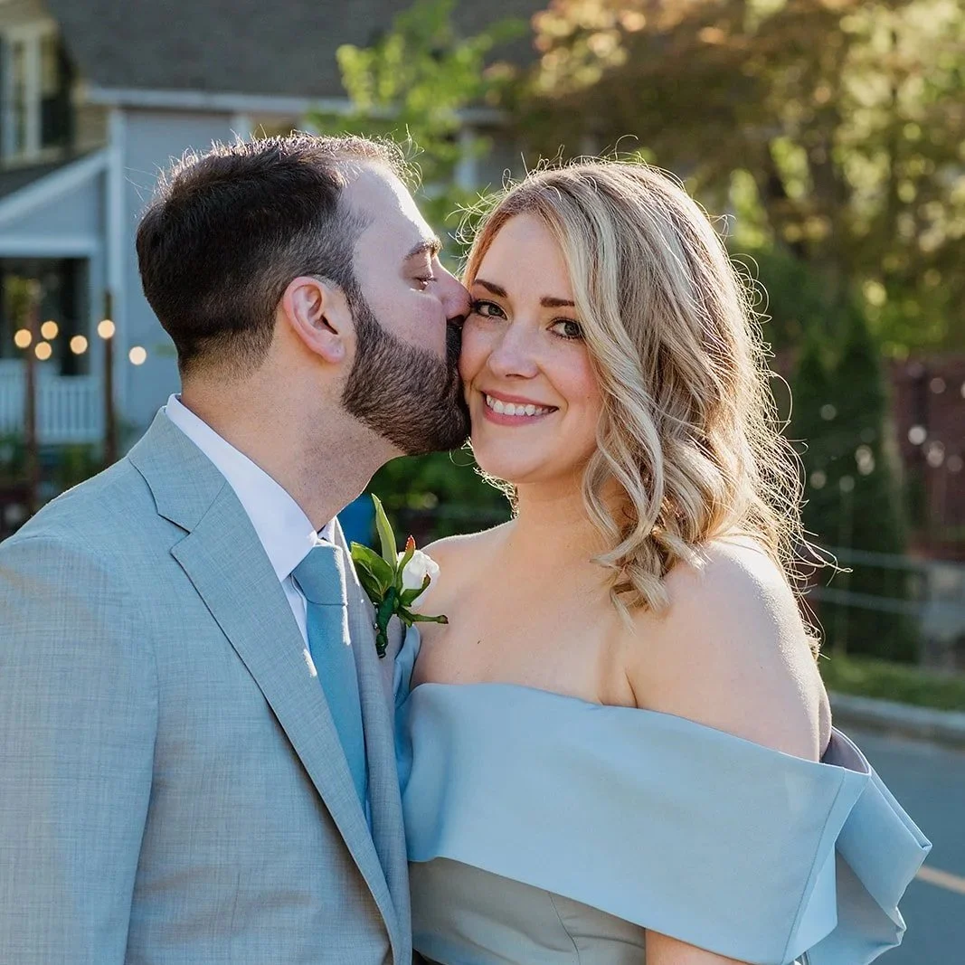 A man in a light gray suit kisses a smiling woman in a strapless light blue dress on her cheek outdoors during daytime.