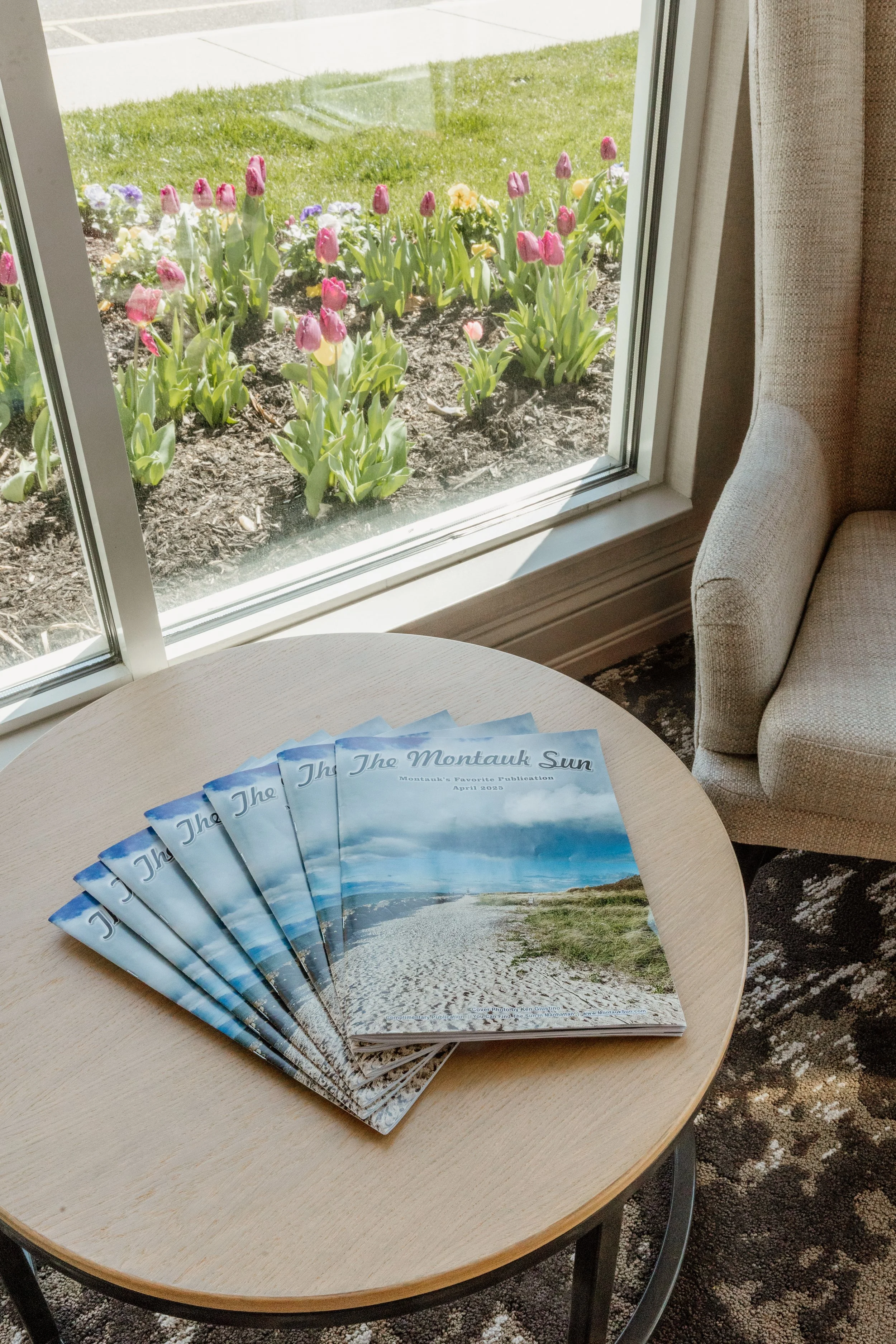 A table with several copies of a magazine titled "The Montauk Sun" featuring a landscape with a sandy path, grassy areas, and a cloudy sky on the cover.