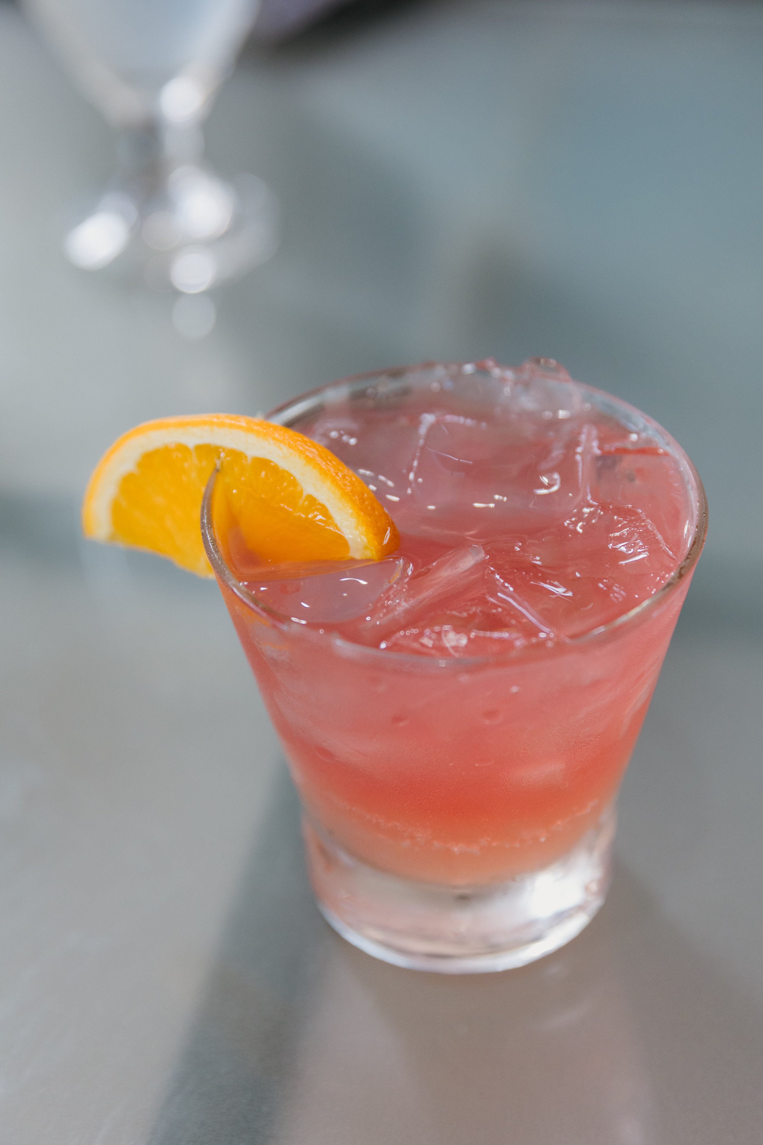 A pink cocktail with ice cubes in a clear glass garnished with an orange wedge, on a gray surface with a blurry glass in the background.