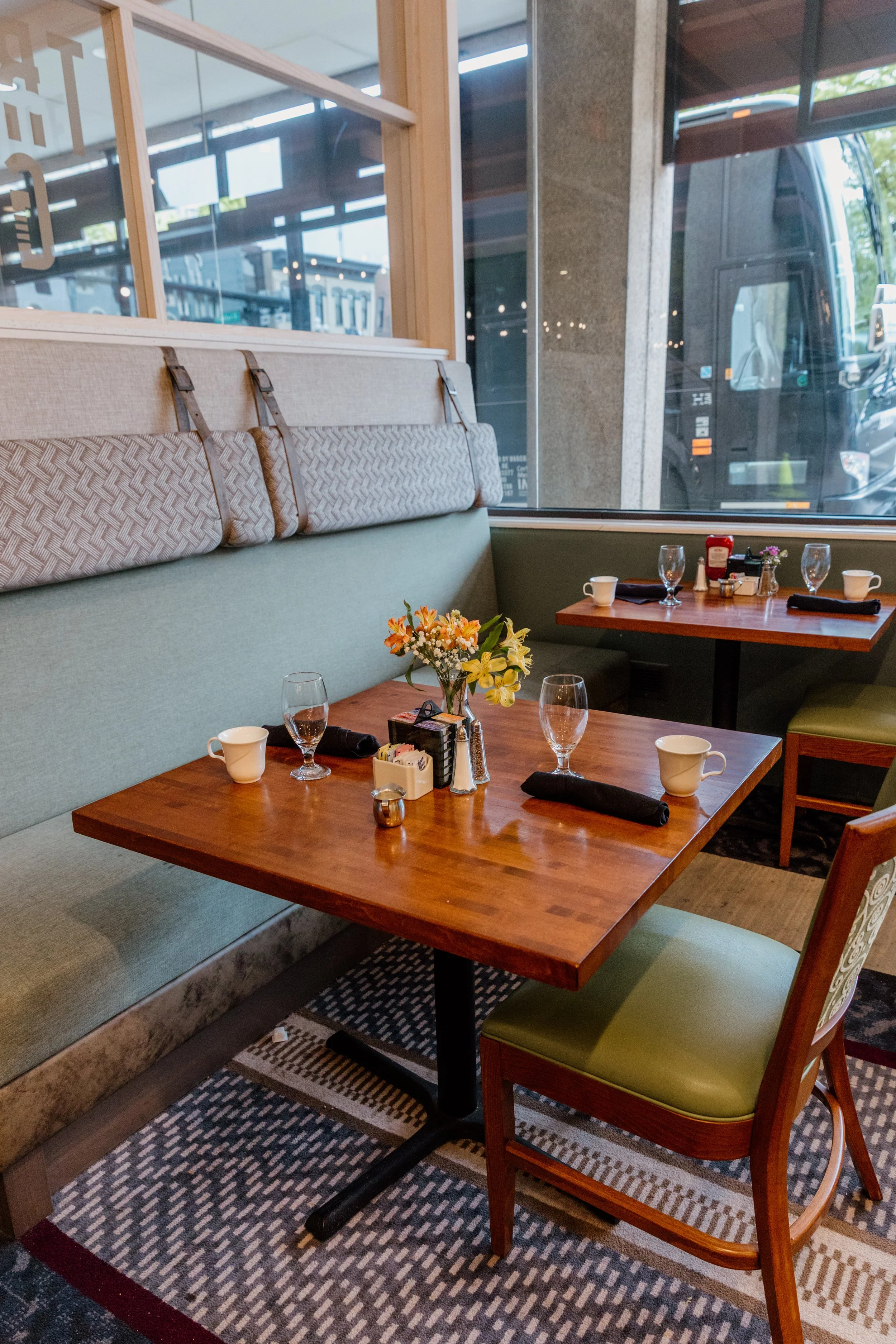 A cozy restaurant booth with a wooden table set for breakfast, featuring cups, water glasses, a flower arrangement, sugar and creamer containers, and black cloth napkins, with a large window showing a sidewalk and bus outside.