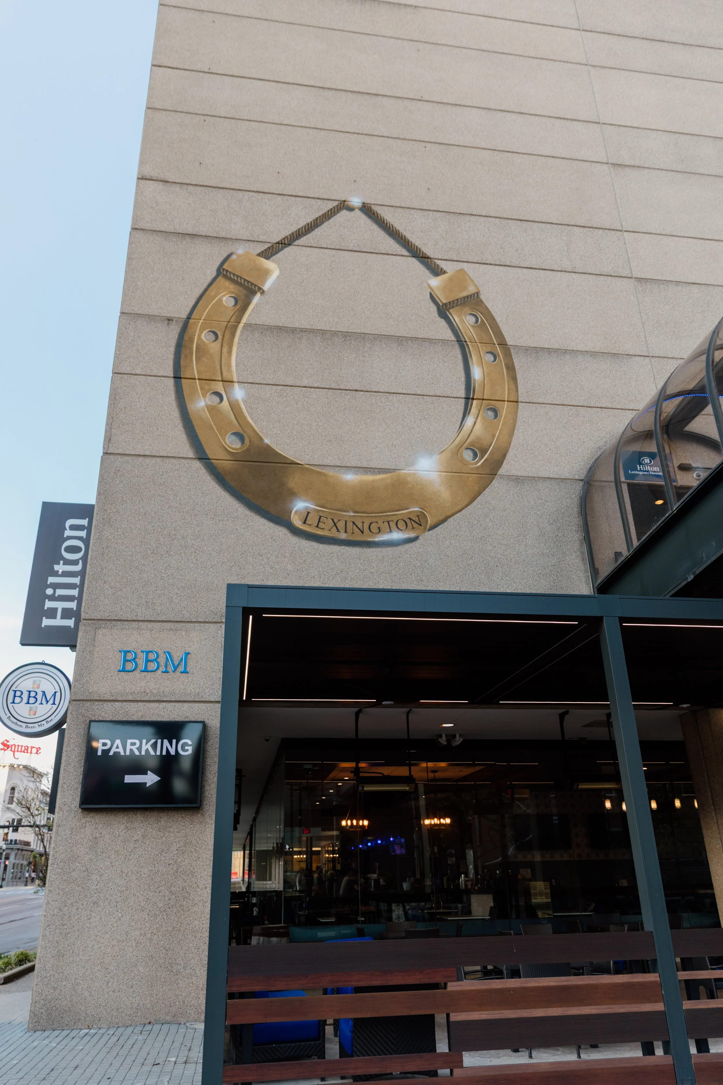 Large decorative horseshoe mounted on exterior wall of building with 'Lexington' inscription, adjacent parking sign pointing to the right, and entrance to a restaurant or bar visible below.