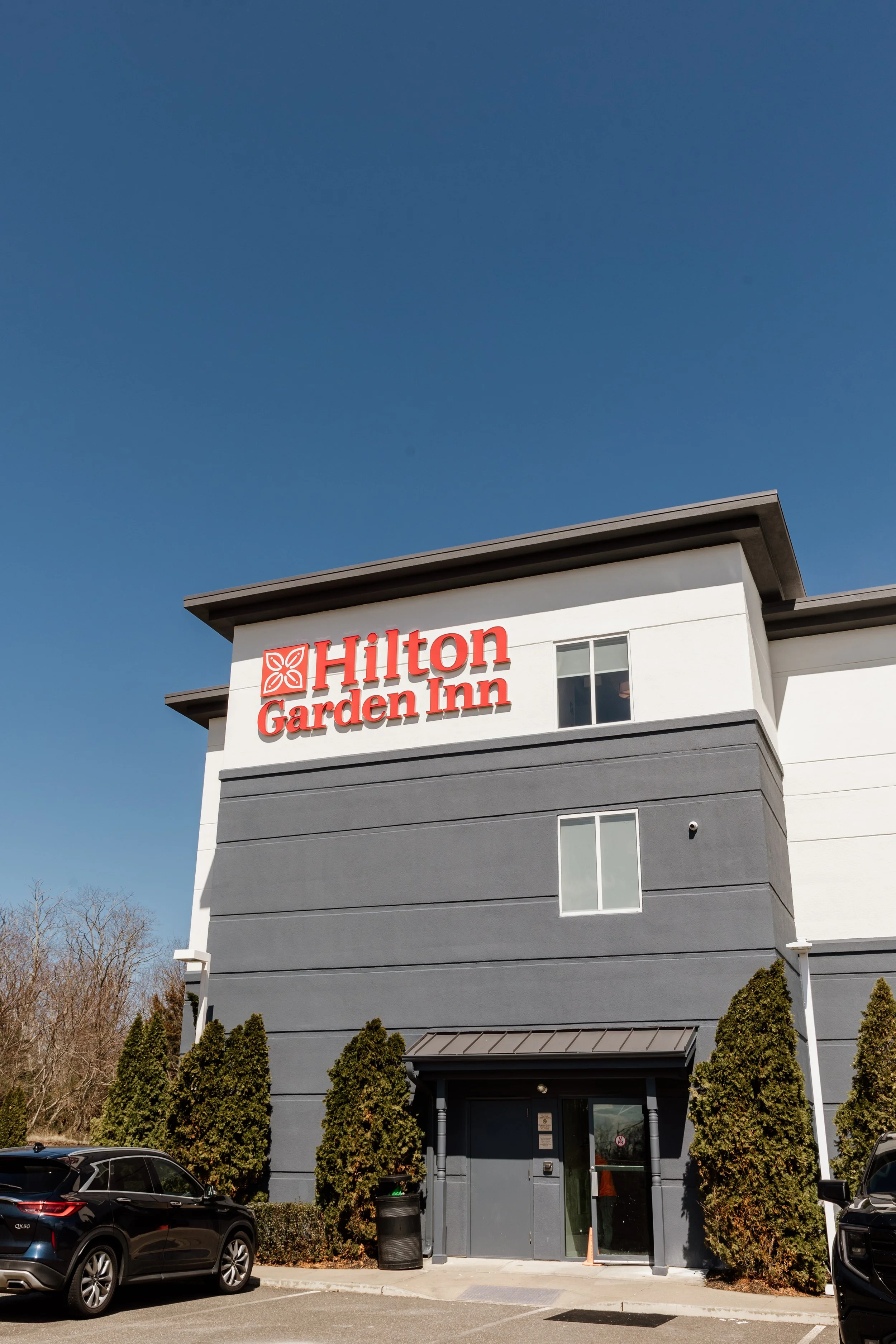Exterior of a Hilton Garden Inn hotel with a large sign, parking lot, and tall bushes in front, under a clear blue sky.