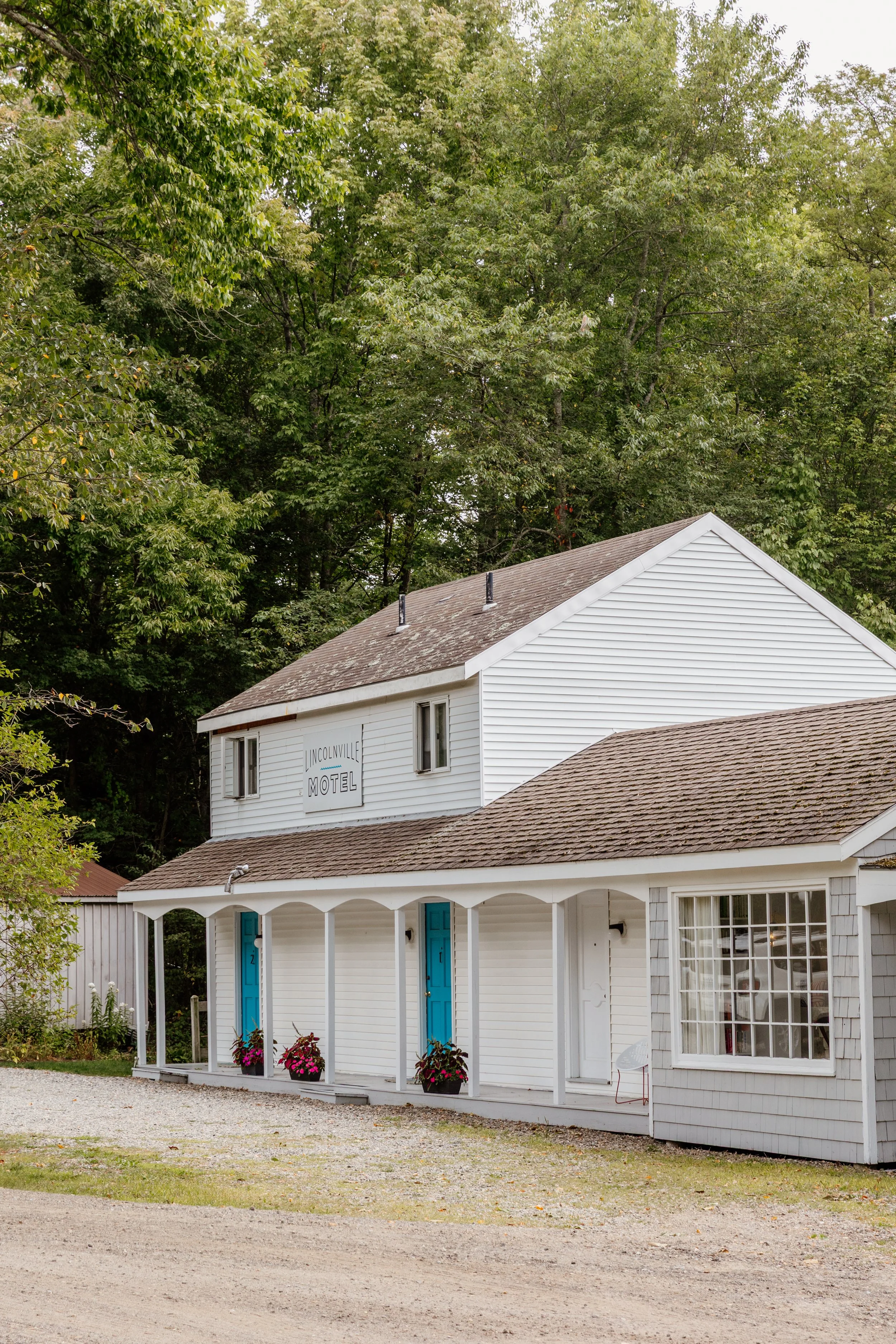 White two-story motel building with blue doors, surrounded by trees, with a gravel parking area in front.