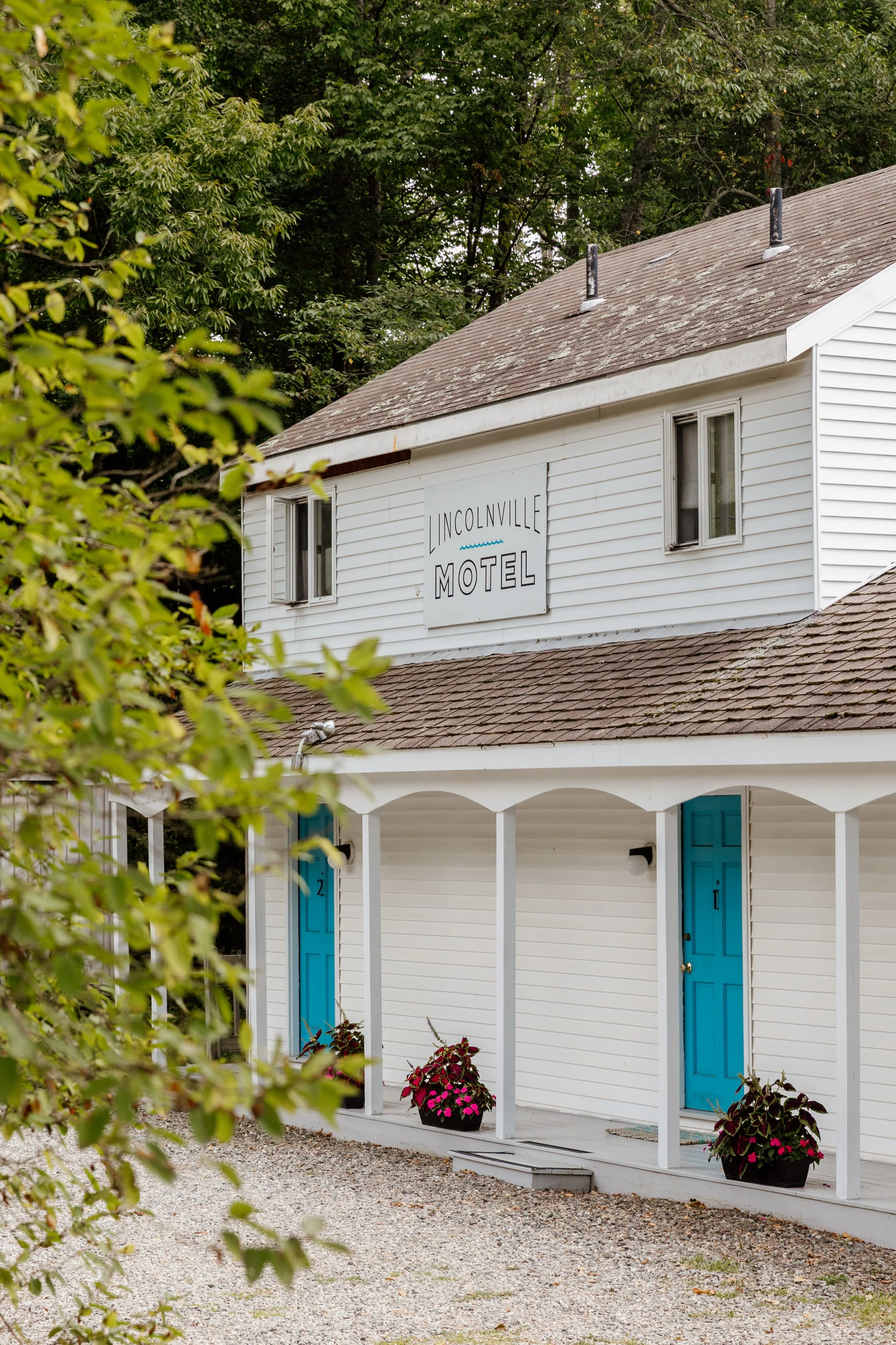 Two-story white building with a sign that reads 'Inconville Motel,' featuring a porch with blue doors, potted plants with flowers, and surrounded by greenery.