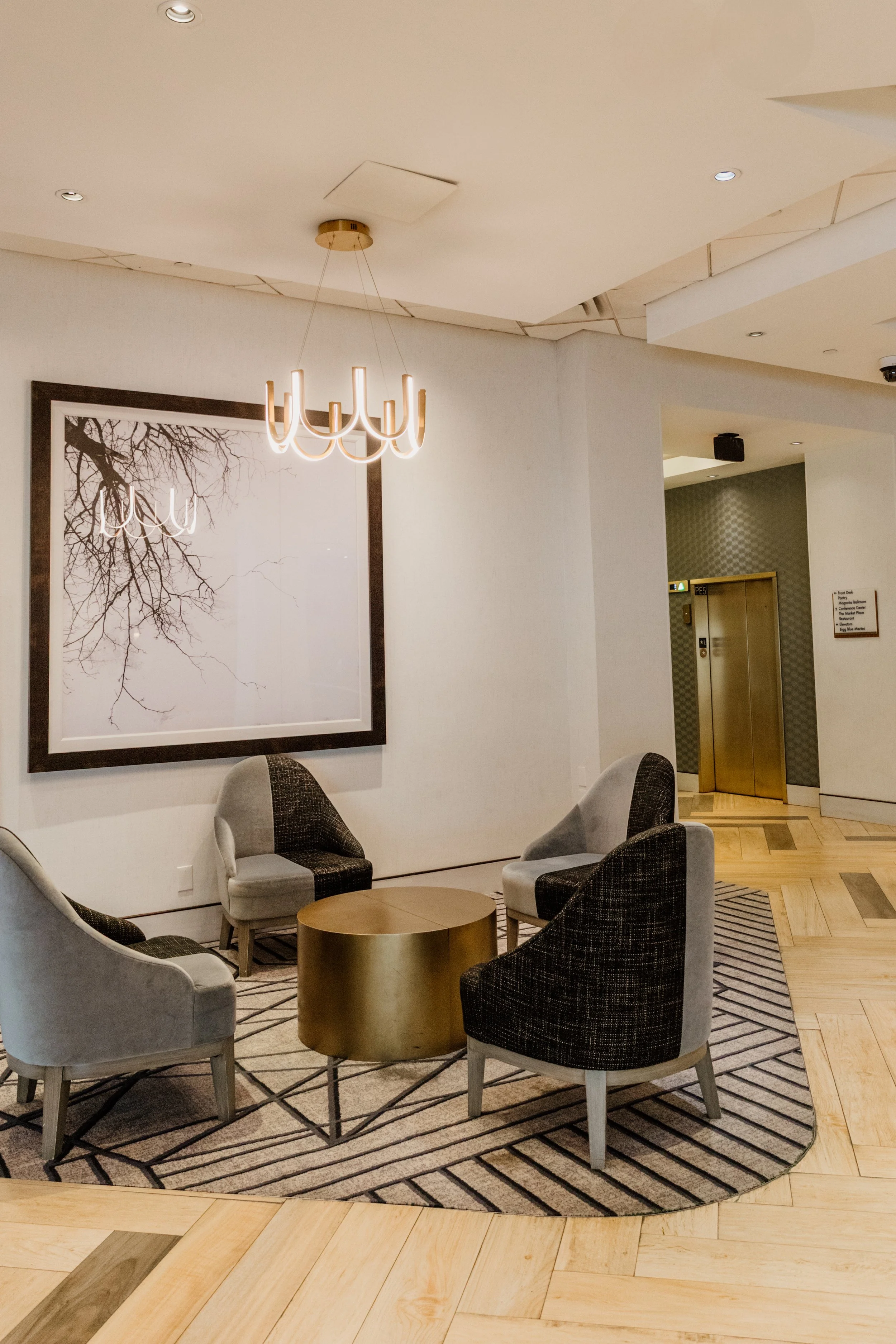 A sleek and sophisticated hotel lobby seating area at the Hilton Lexington Downtown in Lexington, Kentucky, with contemporary furnishings, a gold coffee table, a striking black and white art print, and a glowing chandelier overhead.