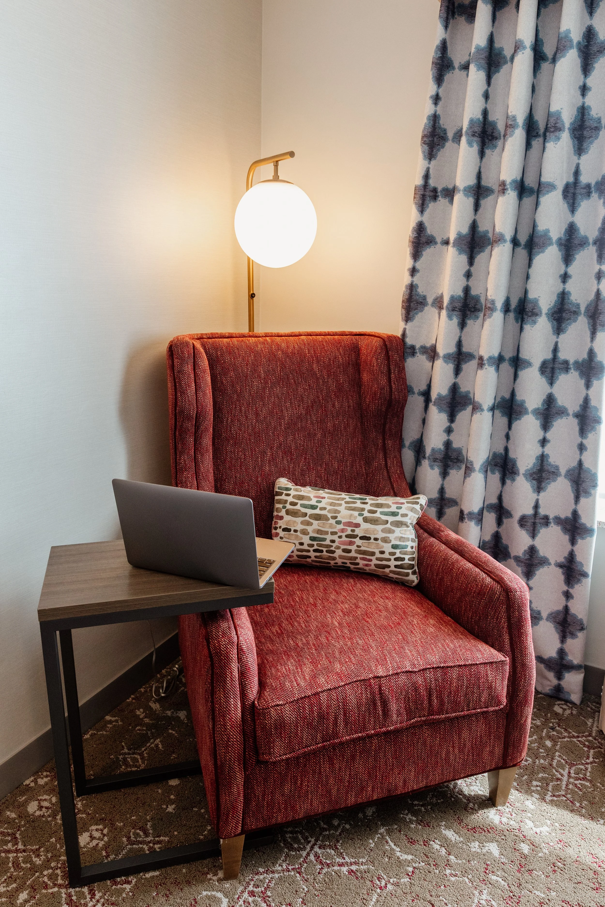 A red upholstered armchair with a decorative pillow, a small wooden side table with a laptop, a wall-mounted spherical lamp, and patterned curtains in a cozy room.
