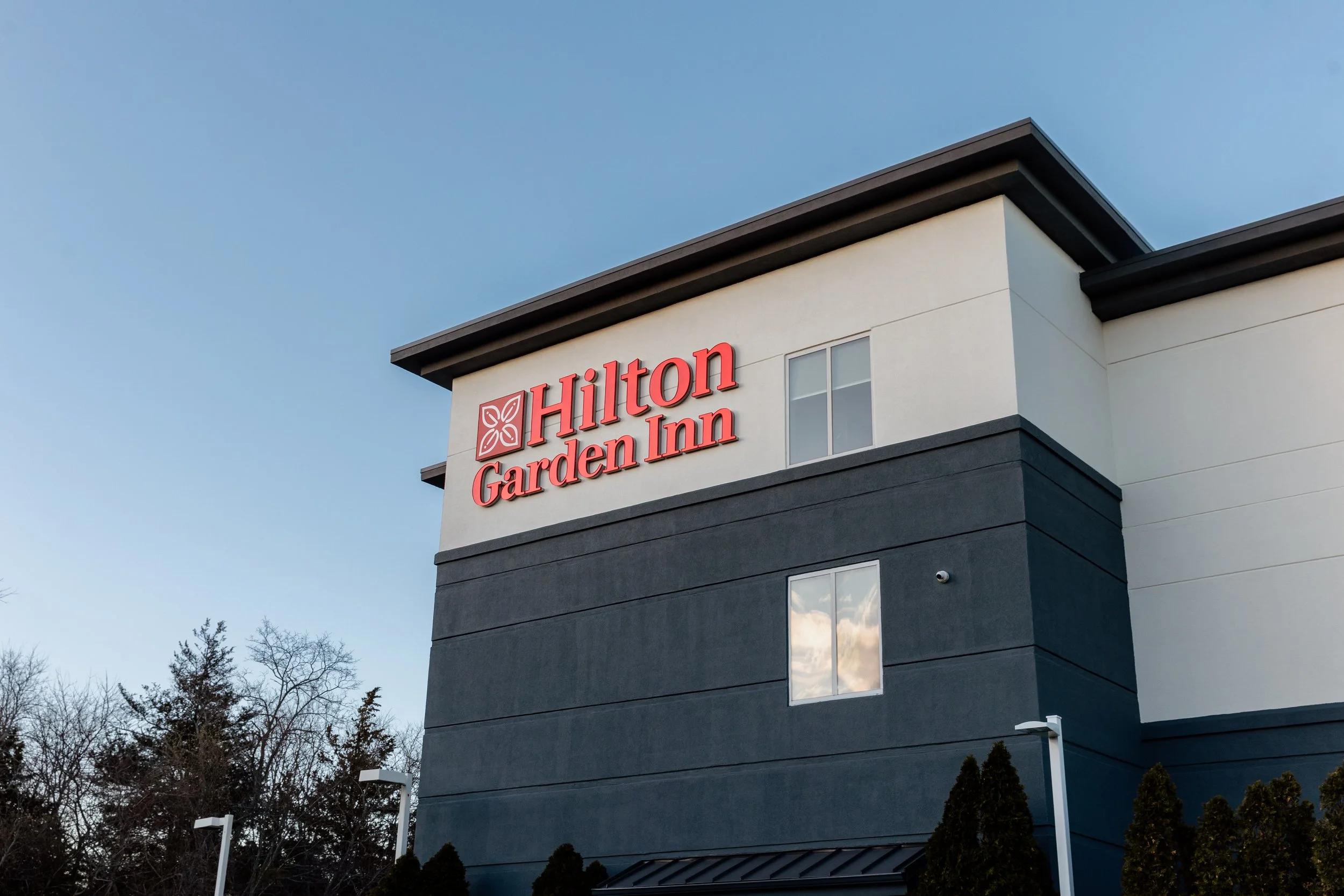 Exterior of a Hilton Garden Inn hotel with the sign visible on the building, trees, and a clear blue sky in the background.