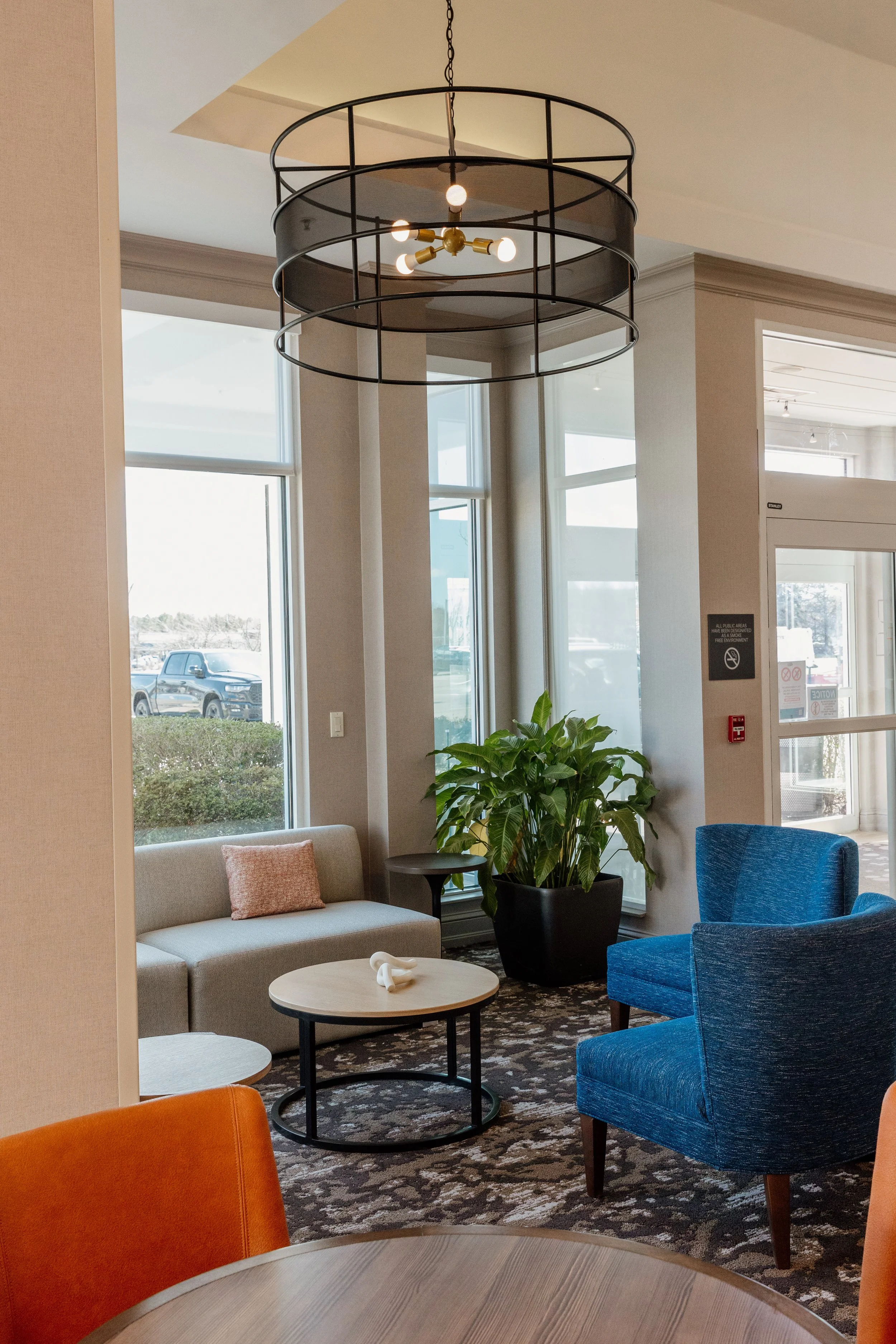 A modern hotel lobby with a round, black metal chandelier, beige sofa with pink pillow, blue armchair, large potted green plant, and round coffee tables, with large windows and a patterned carpet.
