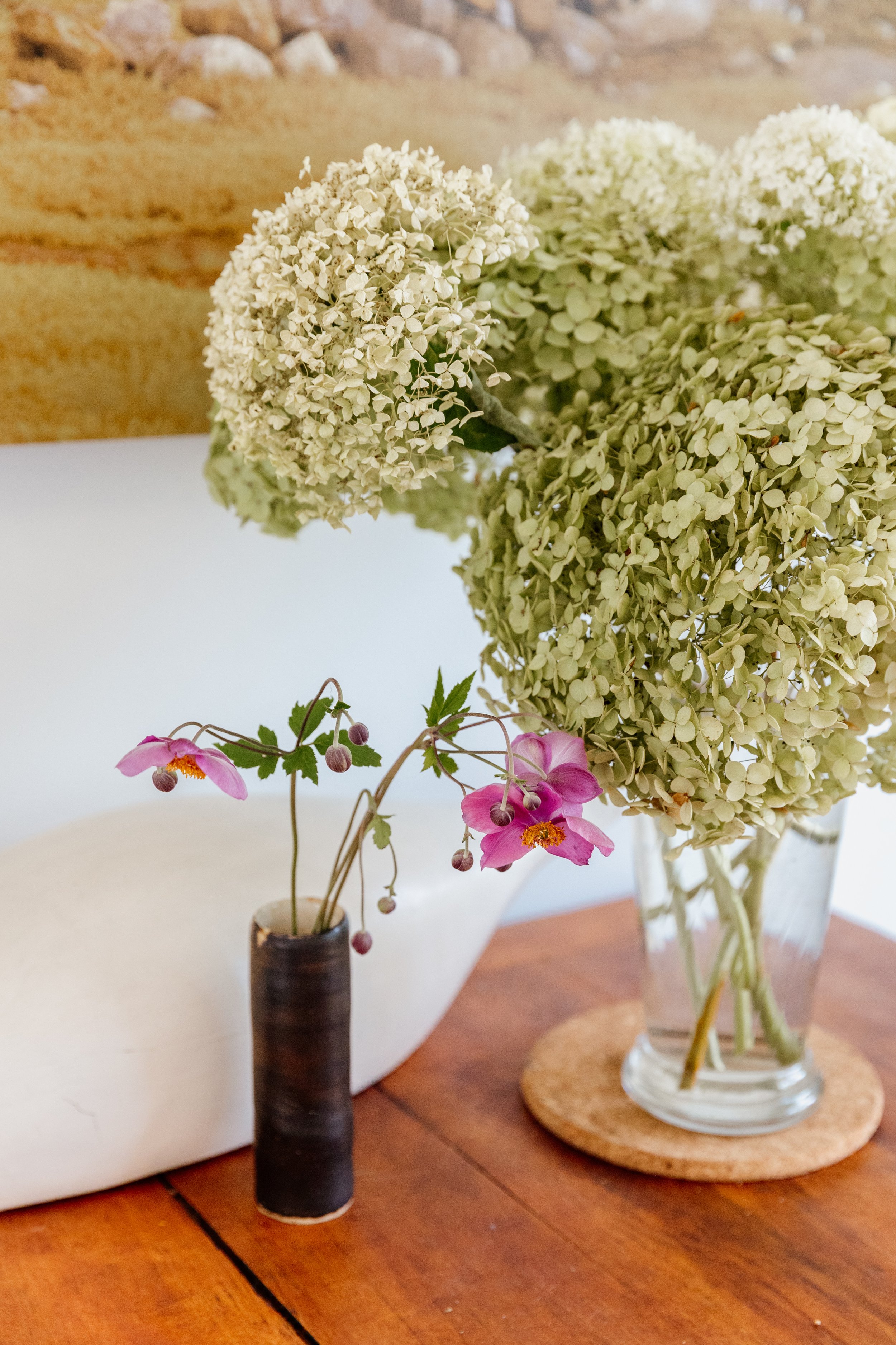 A bouquet of white hydrangeas in a glass vase and a small branch with pink flowers in a dark vase on a wooden table.