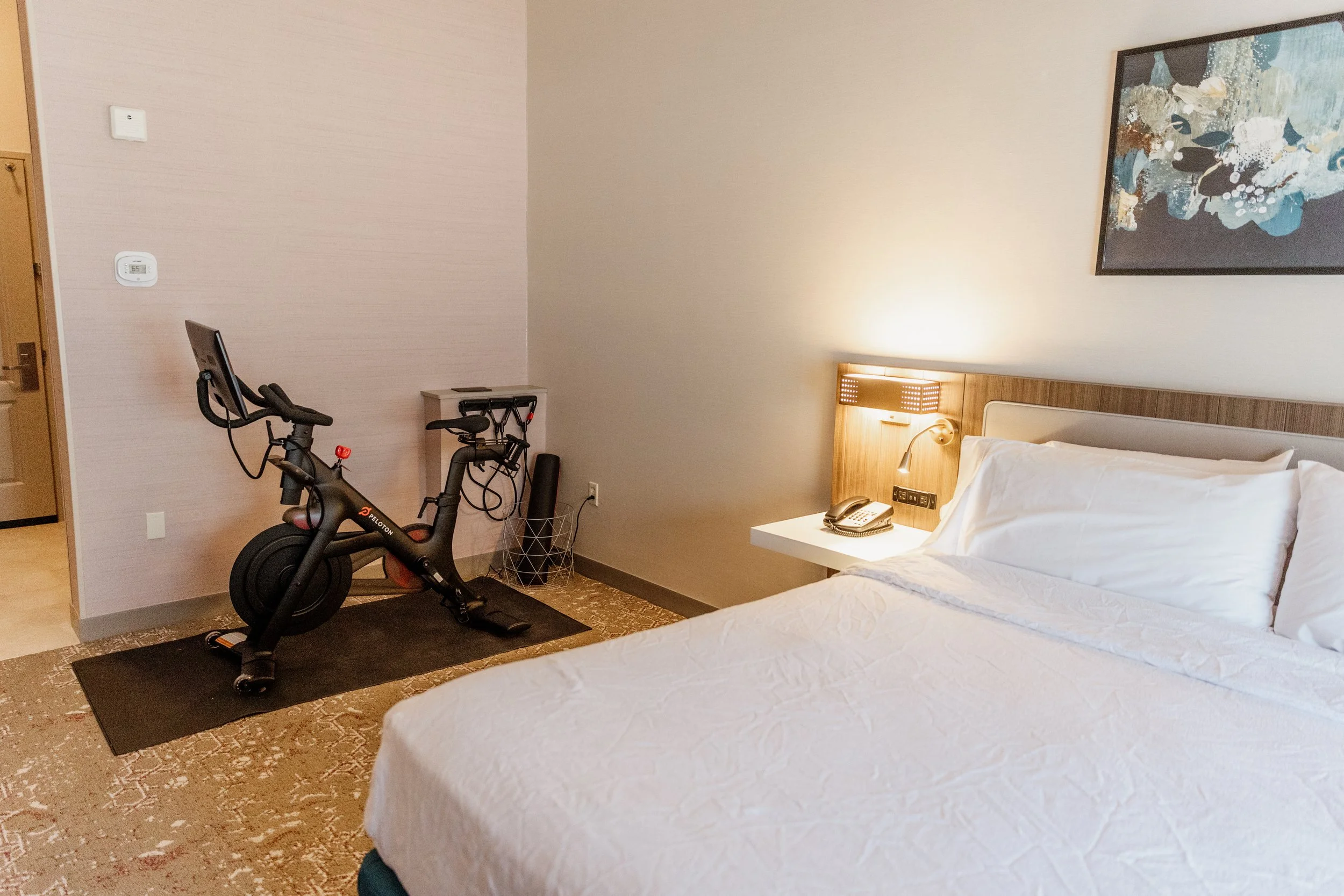 A well-appointed guest room at the Hilton Garden Inn in Riverhead, New York, complete with a cozy bed, warm lighting, and an in-room Peloton bike for a little extra motivation.