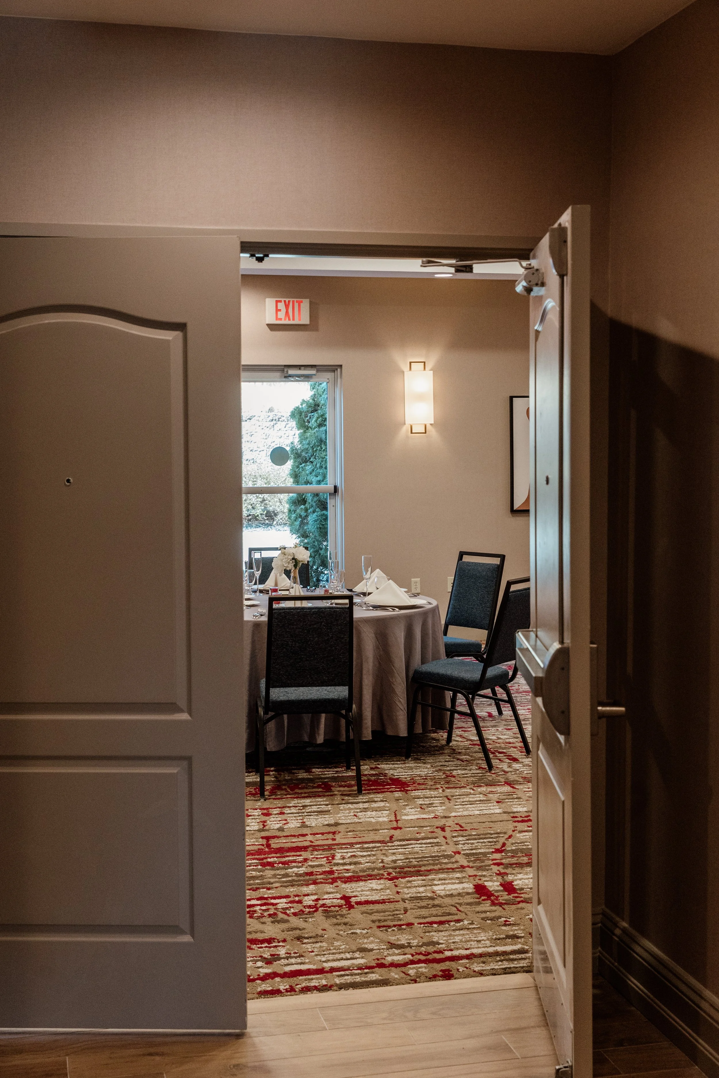 A round banquet table set for a formal event, viewed through an open door, with napkins folded on plates, wine glasses, and a floral centerpiece, in a room with beige walls, a window, and modern light fixtures.