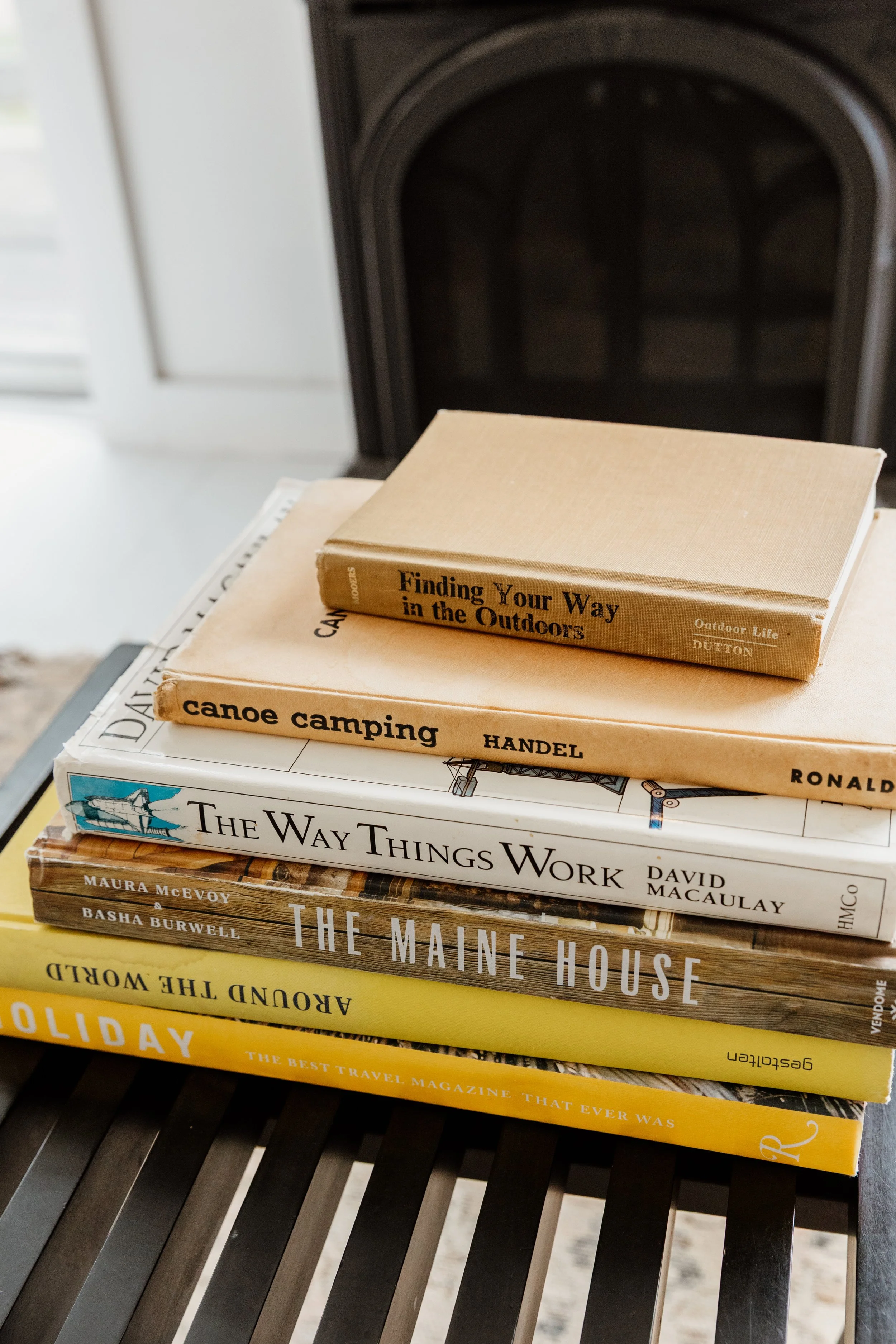 Stack of travel and outdoor-related books on a black slatted table, with a fireplace in the background.