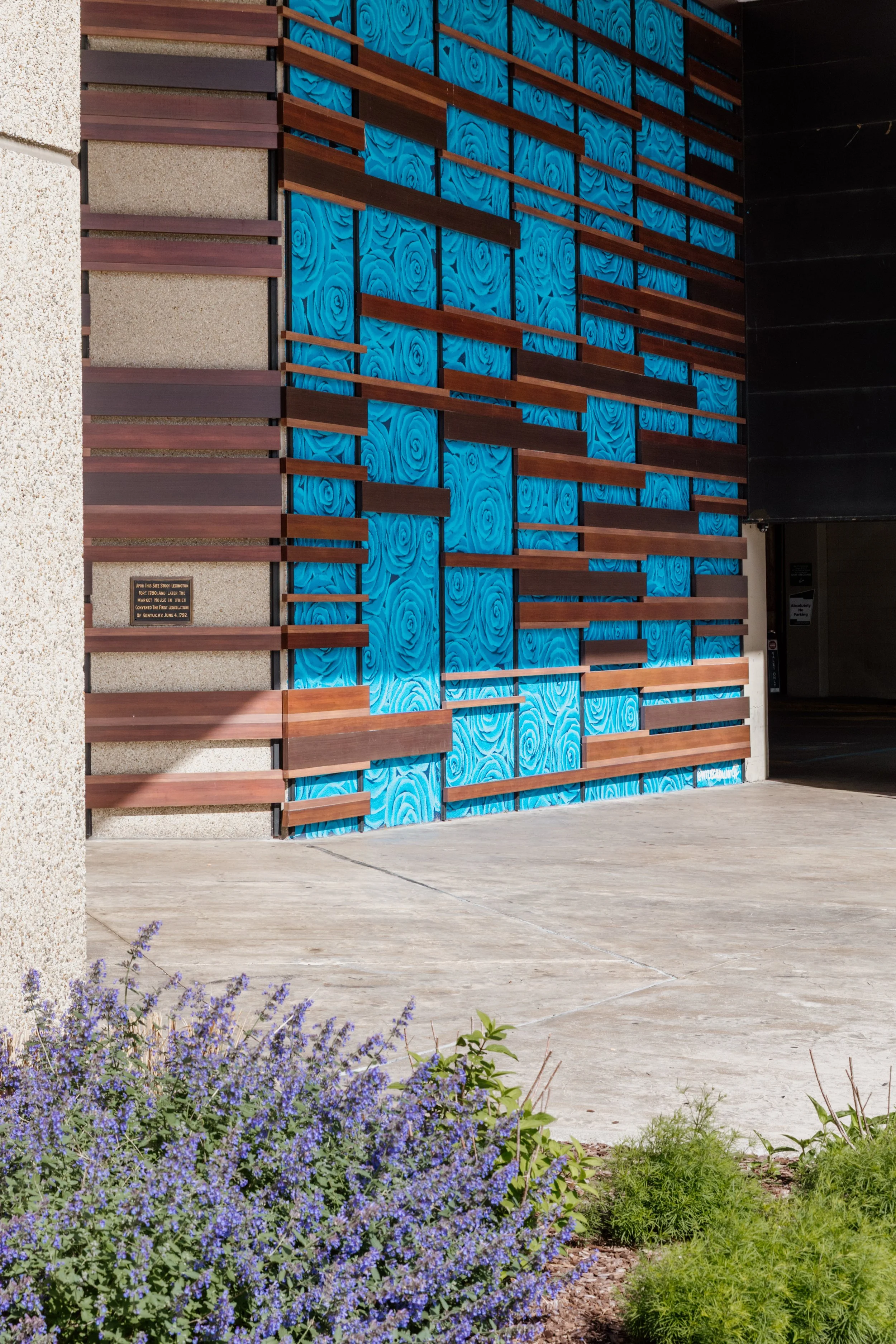 Modern building with a decorative wall featuring blue panels with a rose pattern and horizontal wooden slats, surrounded by a sidewalk and purple flowering plants.