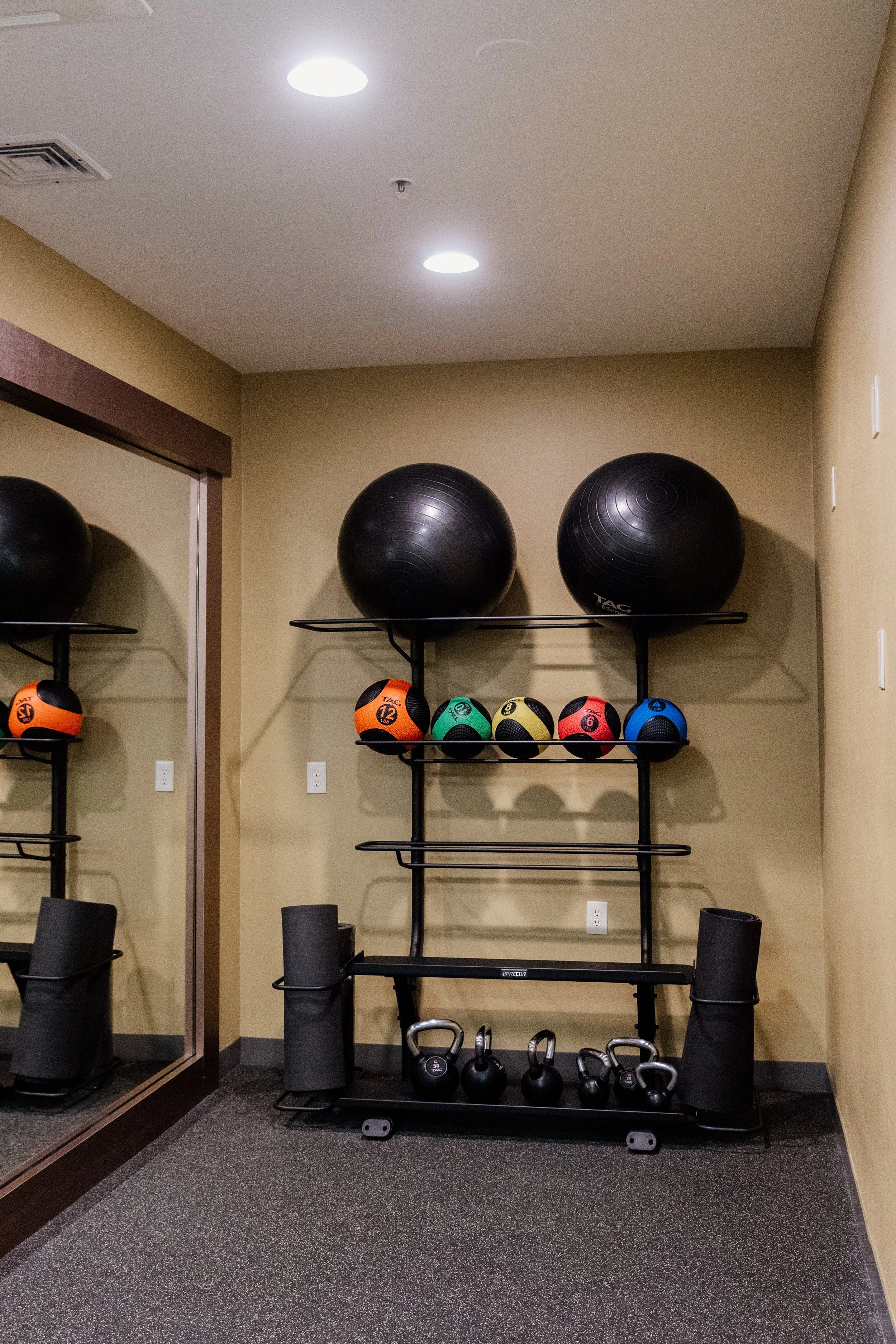 A shelf holding large black exercise balls, smaller medicine balls in orange, green, yellow, red, and blue, and kettlebells, with a mirror to the left reflecting part of the gym equipment.