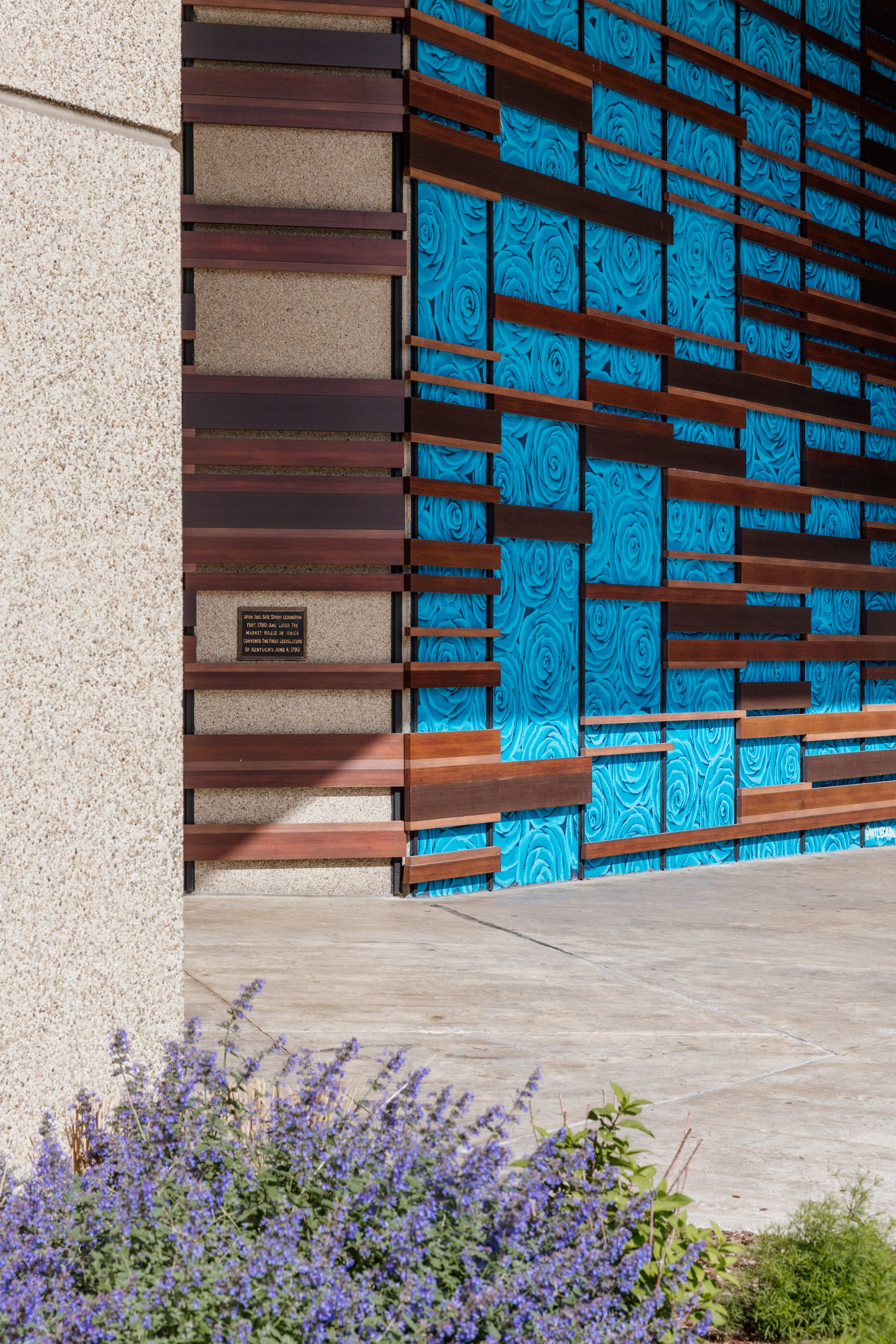 Part of a building exterior with a textured beige wall on the left, a modern design featuring horizontal wooden slats and blue panels with a rose pattern on the right, and purple flowering bush at the bottom.