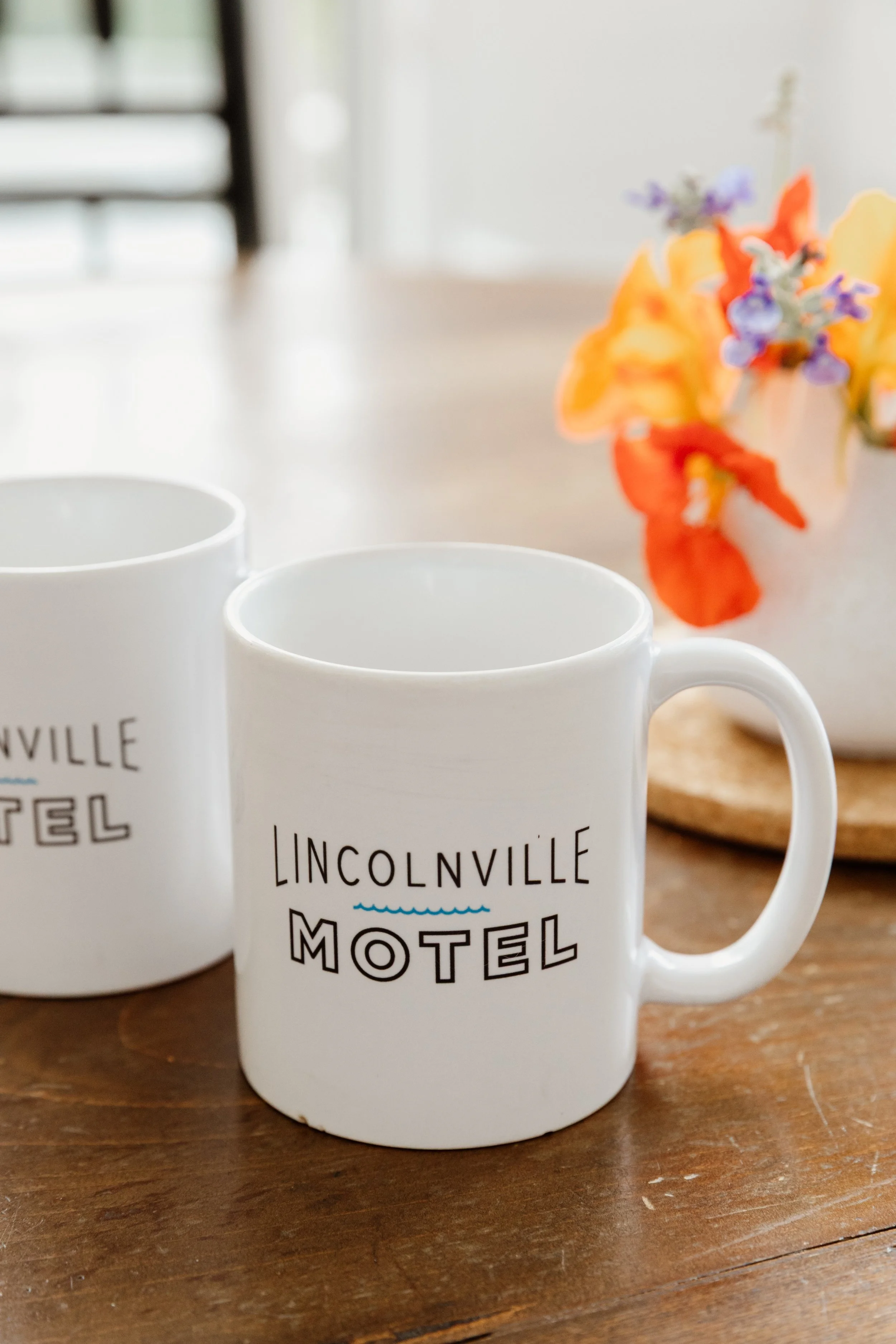 White ceramic mug with 'Lincolnville Motel' printed on it, placed on a wooden table alongside a similar mug and a white vase with colorful flowers.