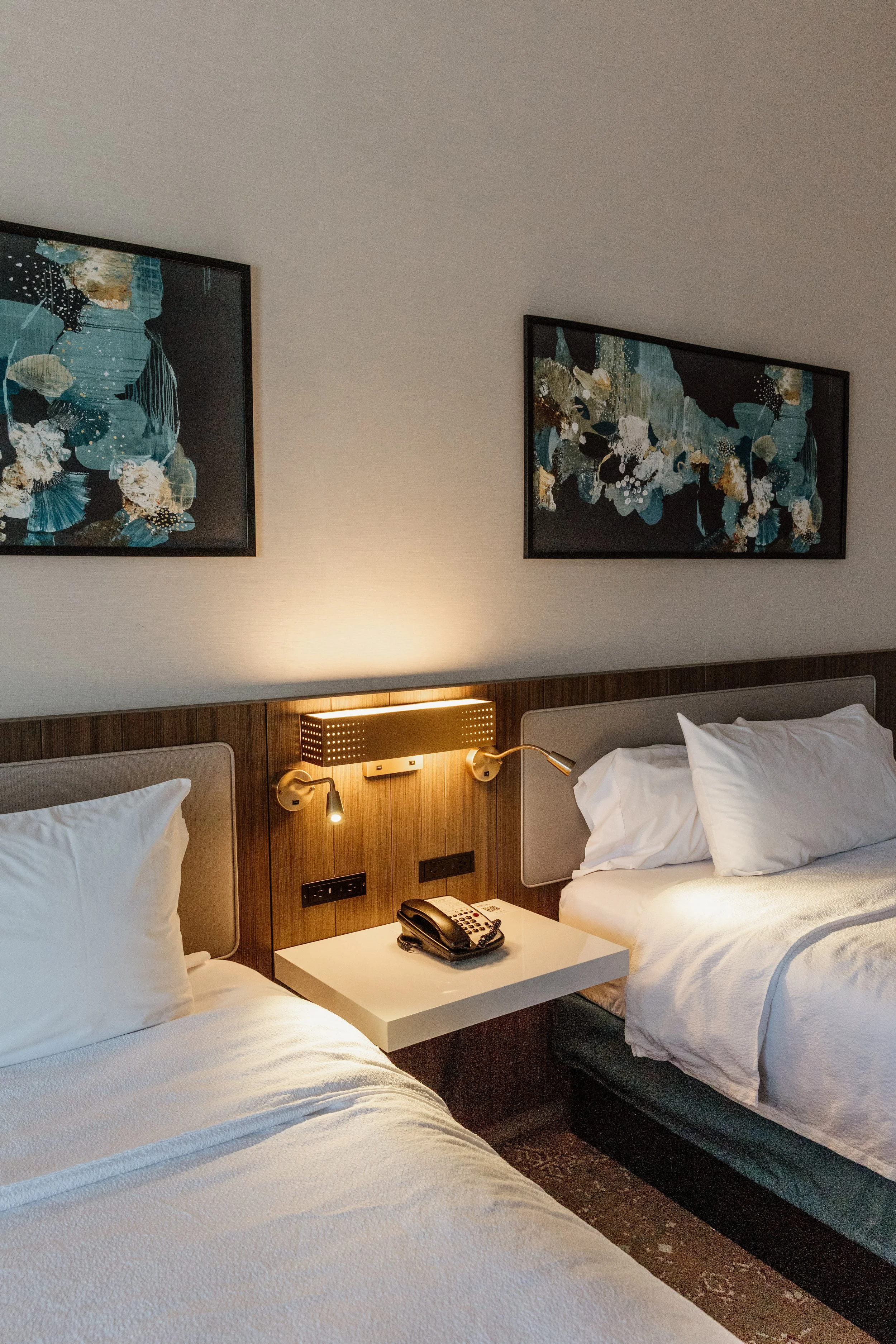Hotel room with two beds, a nightstand, wall-mounted light fixtures, a telephone, and framed artwork on the wall.