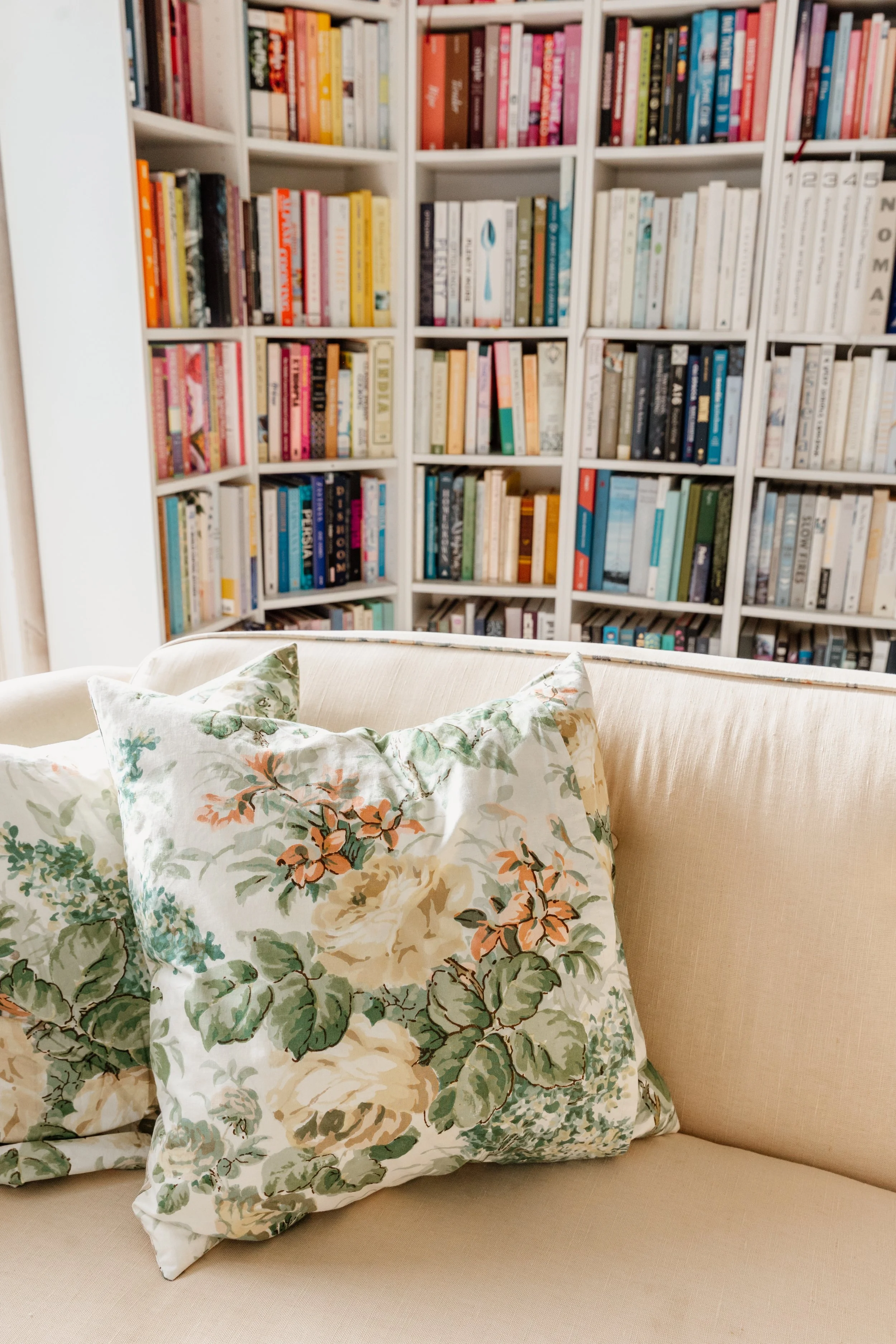 A beige sofa with floral pillows in front of a white bookshelf filled with a variety of books.