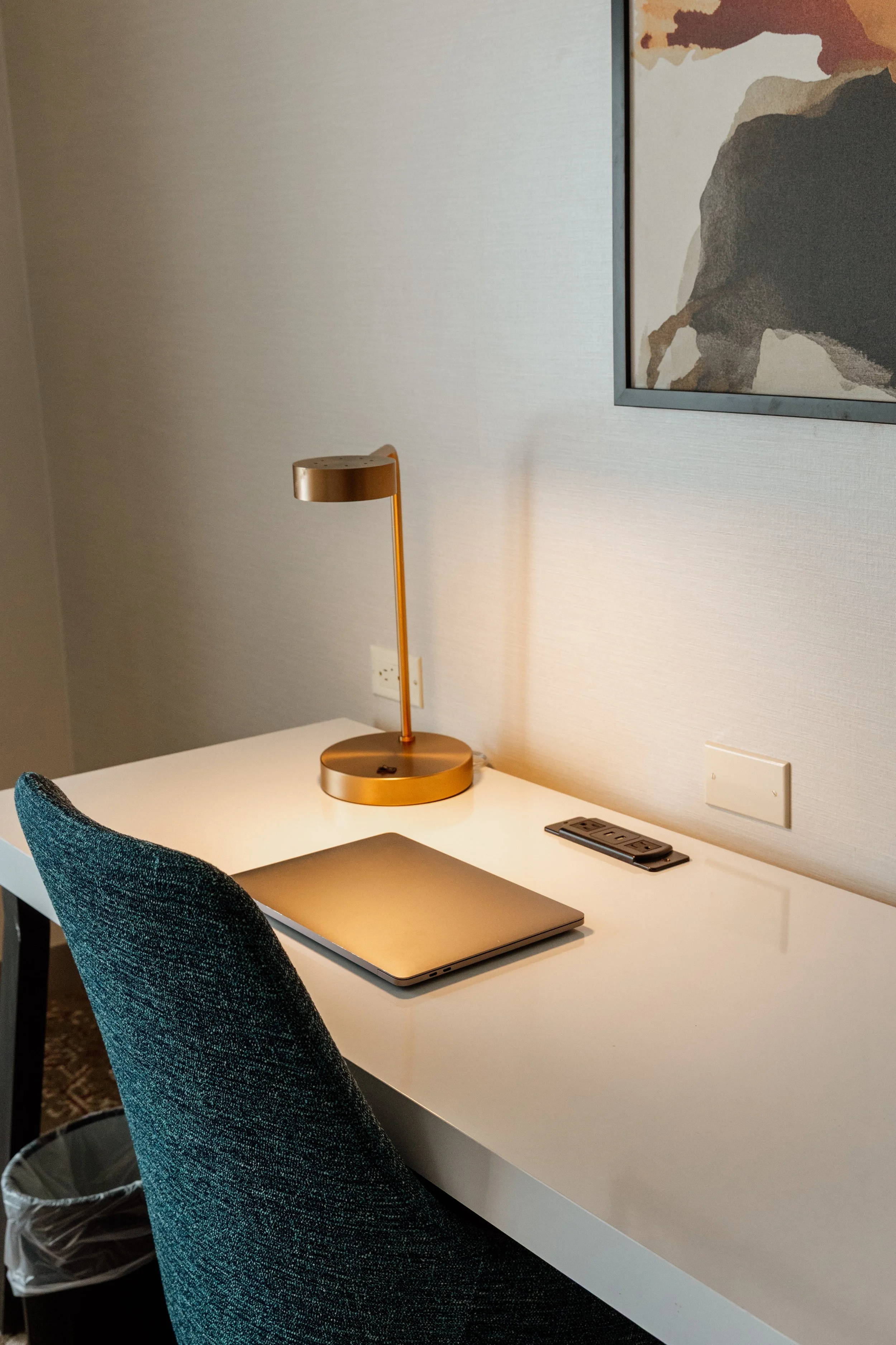 A photo of a white desk with a closed laptop, a desk lamp, and a remote control. There is a blue upholstered chair and a wall with a framed art piece and an outlet with cables.