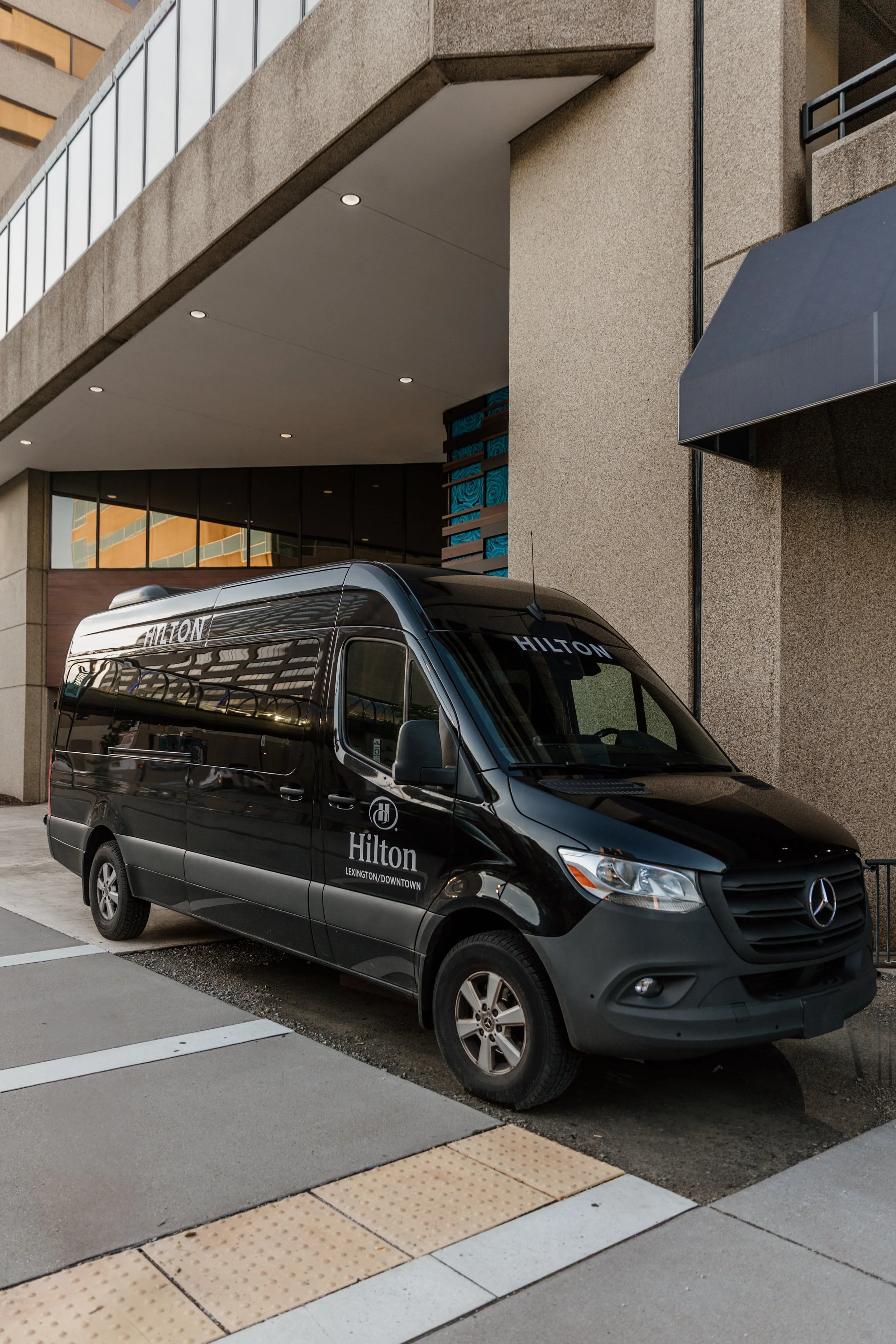 Black Mercedes-Benz van parked on city street with Hilton hotel branding on the side.