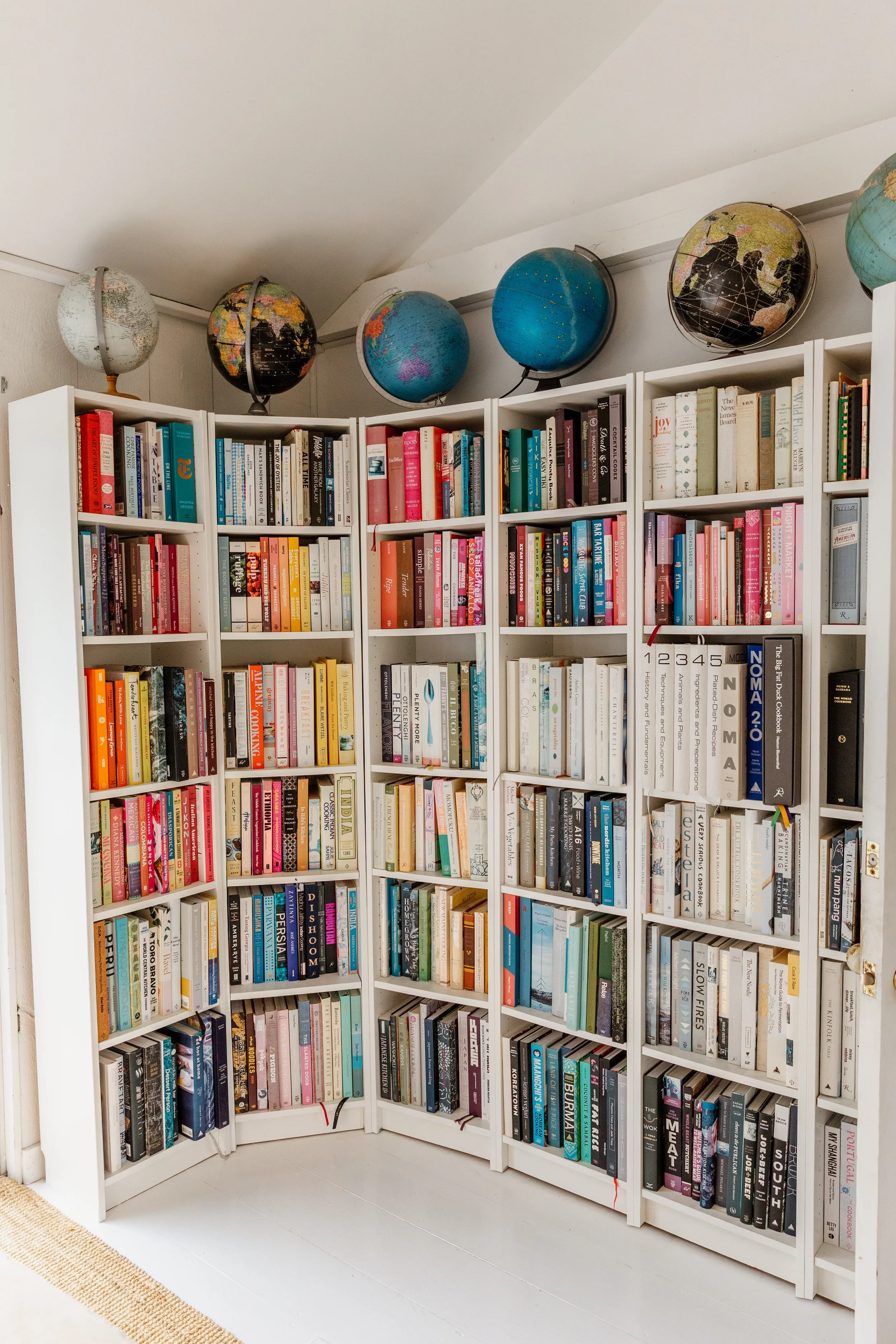 A white bookshelf filled with books, topped with globes, located in a room with white walls and a white floor.