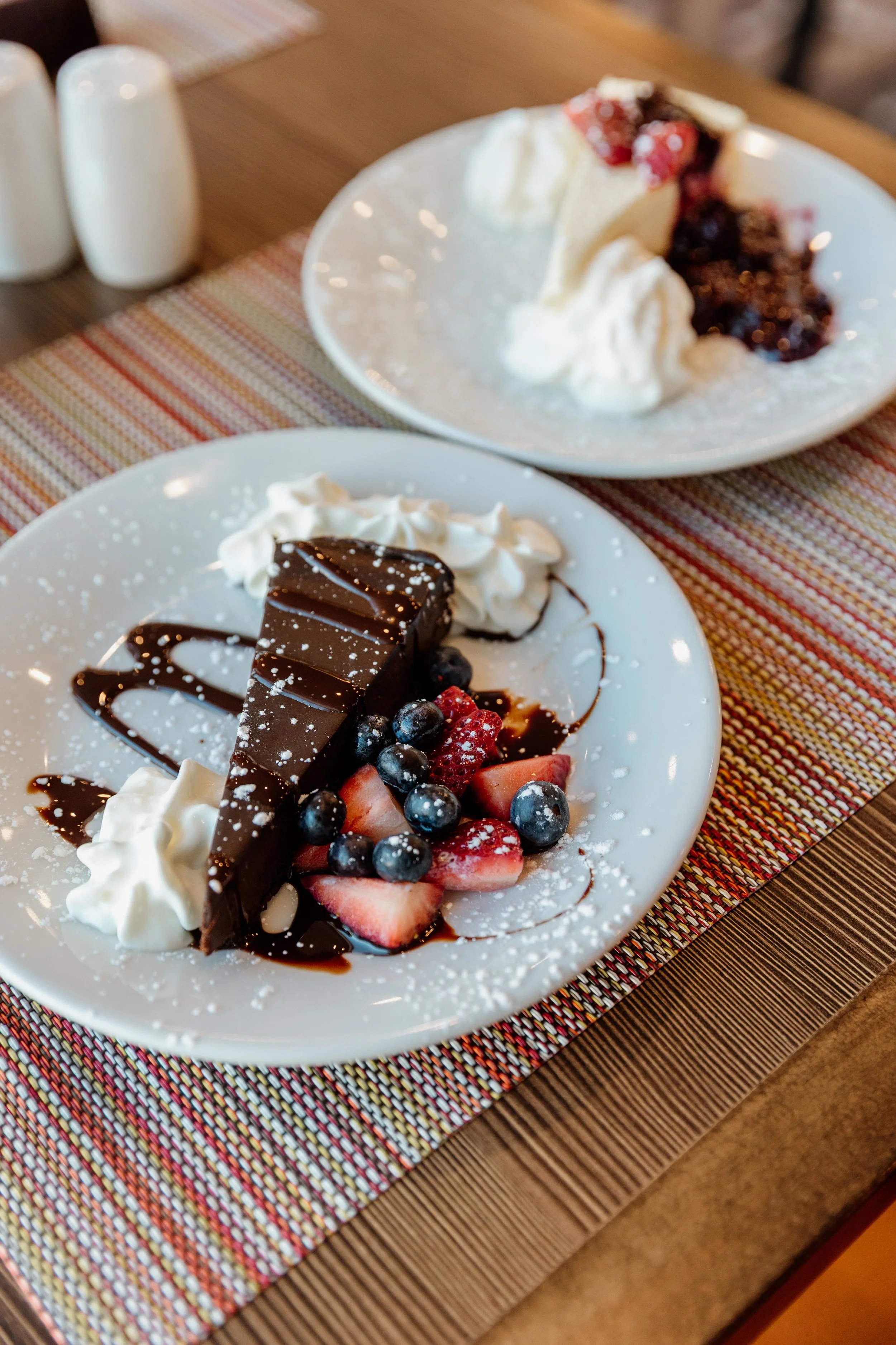 Plate of chocolate mousse cake garnished with strawberries, blueberries, whipped cream, drizzled chocolate sauce, and powdered sugar, with a dessert with berry topping and whipped cream in the background.
