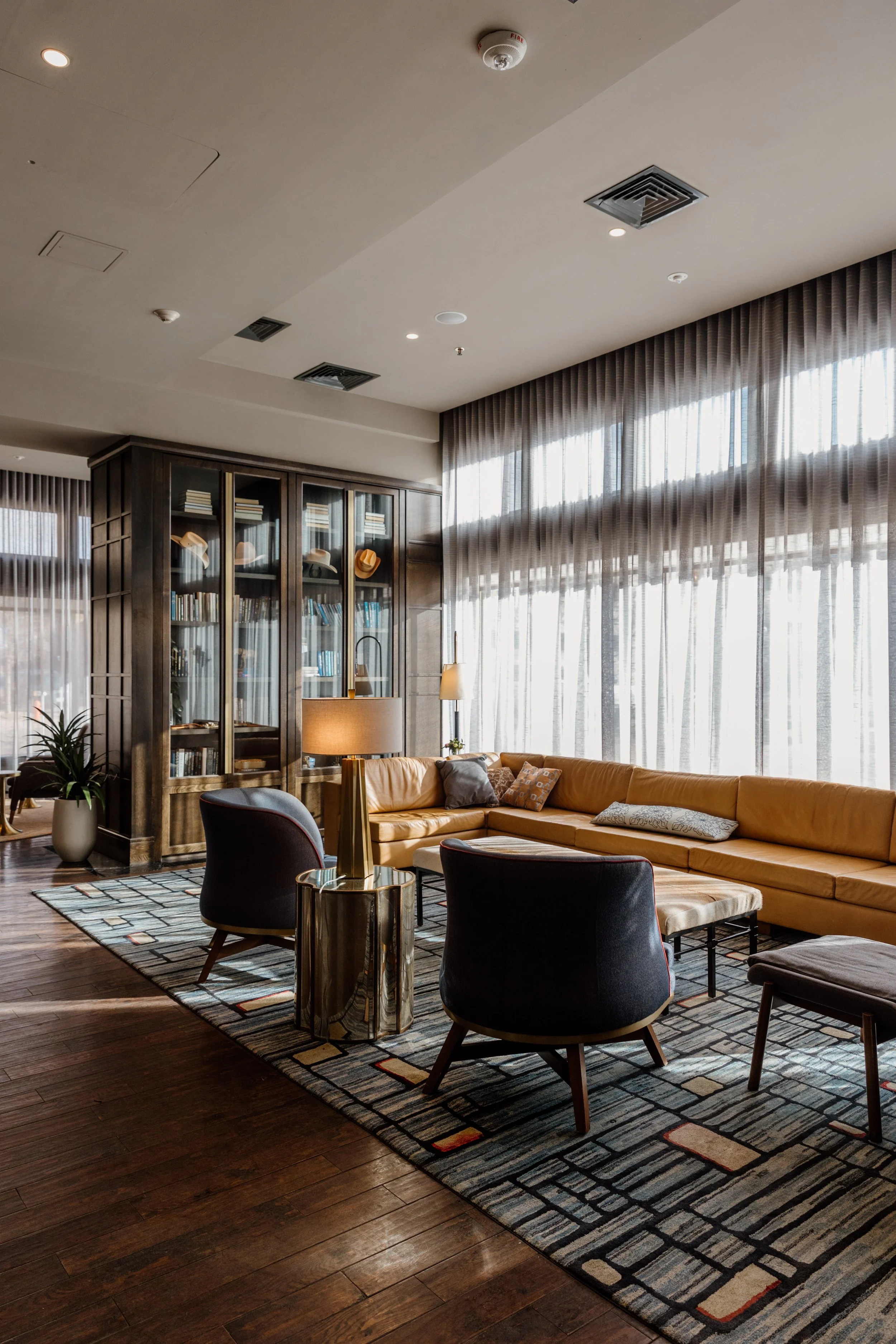 Modern hotel lobby with large windows, wood flooring, a beige sectional sofa, black chairs, a glass table, and a built-in bookshelf display with hats.