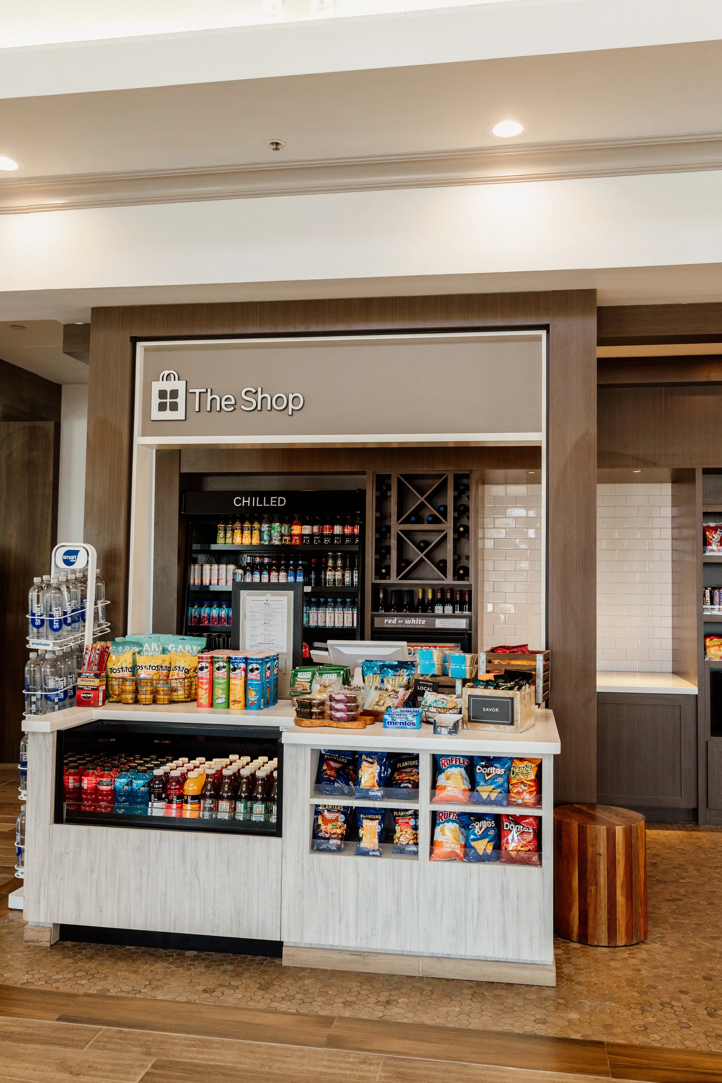 A small retail kiosk in a hotel or cafe, featuring snacks, bottled drinks, and a refrigerator, labeled 'The Shop'.