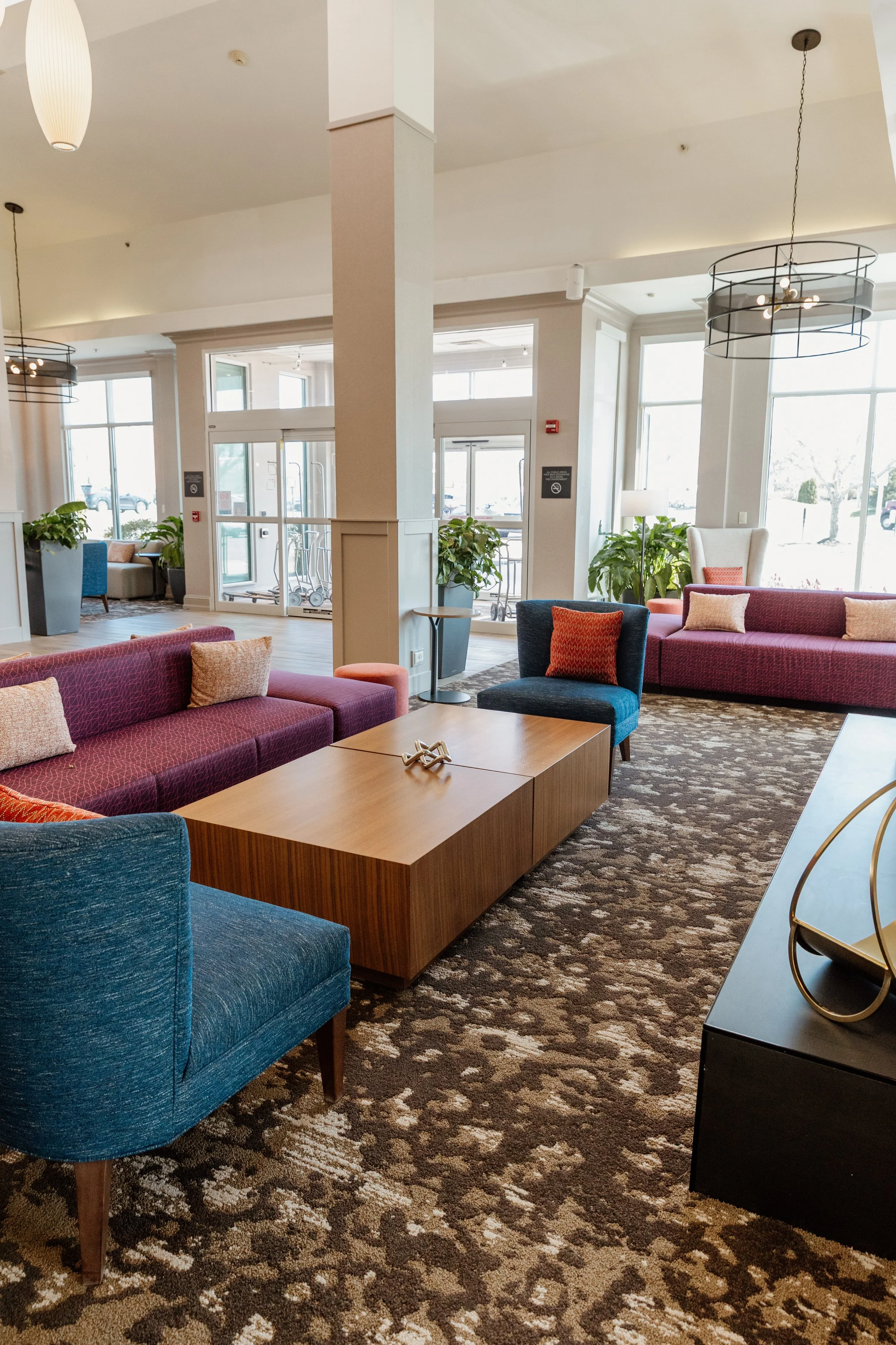 A bright and welcoming hotel lobby at the Hilton Garden Inn in Riverhead, New York, with colorful sofas, a wooden coffee table, cozy armchairs, and plenty of natural light pouring through large windows.