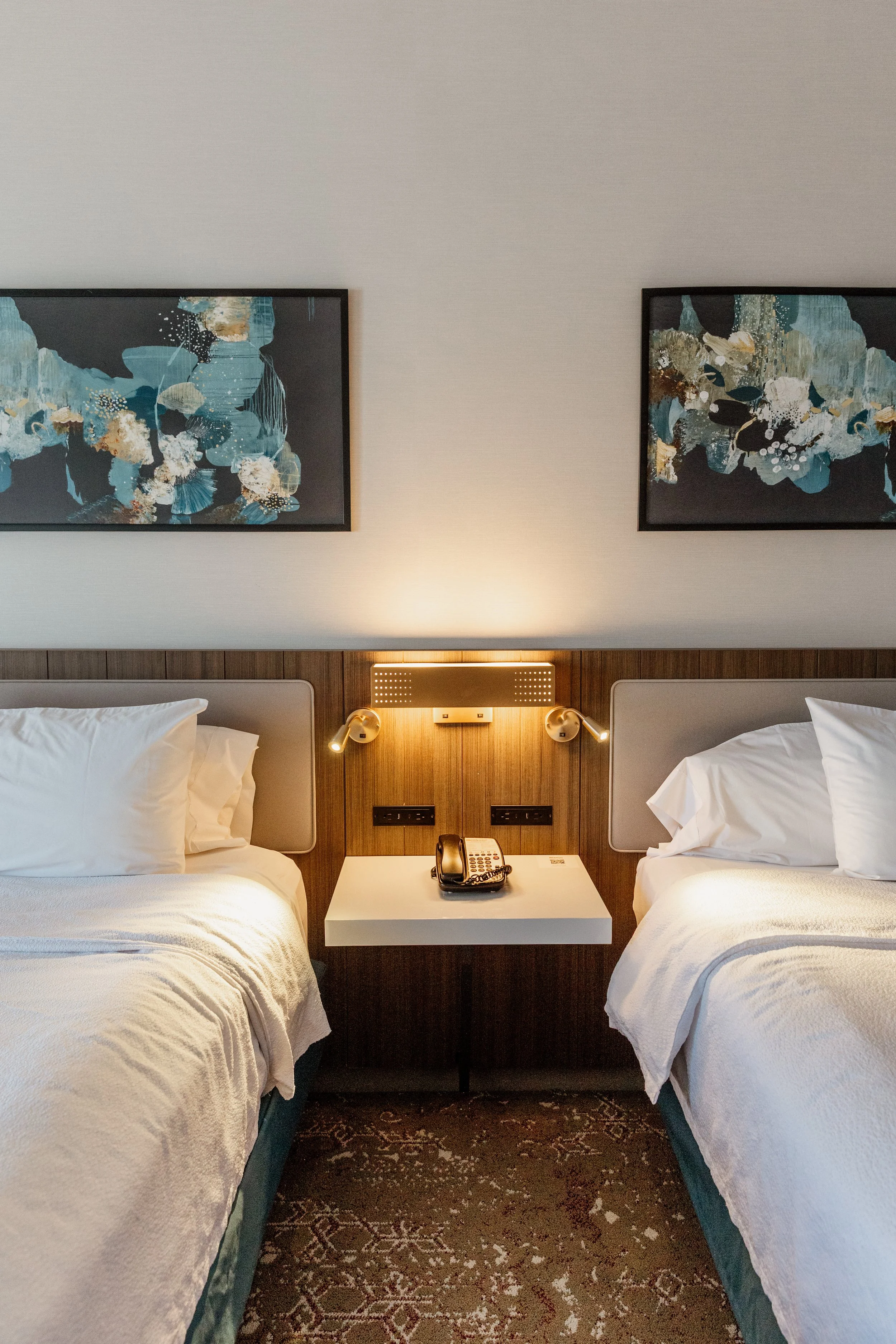 Hotel room with two beds, a nightstand with a telephone, wall-mounted lamps, and two abstract paintings above the beds.