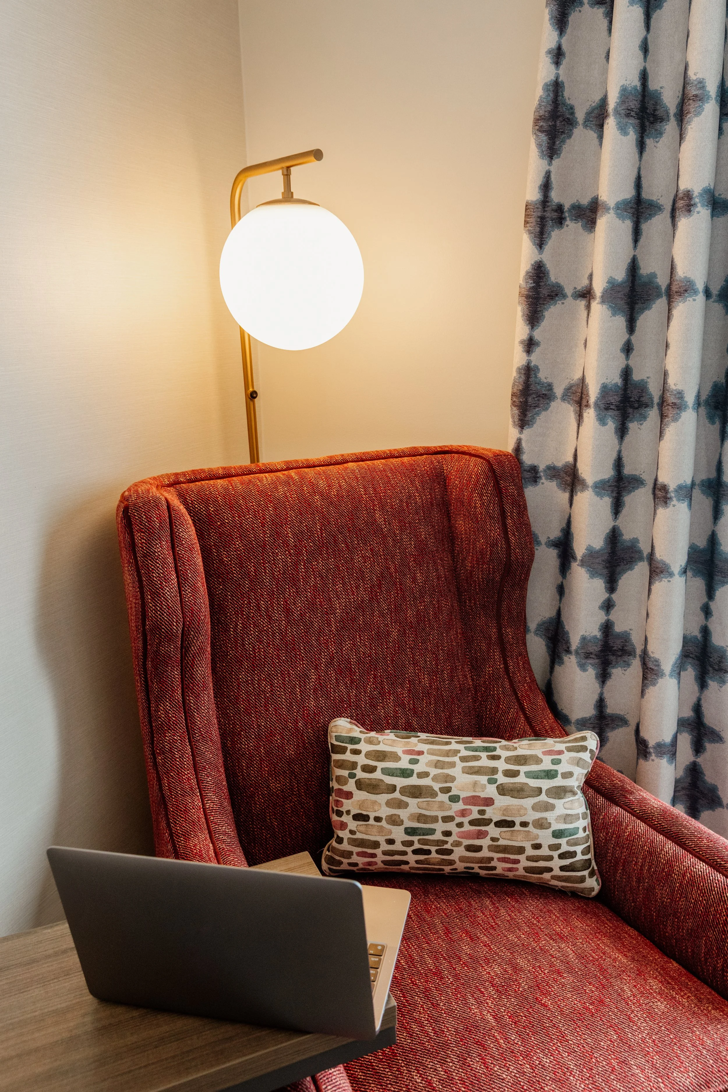 A red upholstered armchair with a patterned rectangular pillow, a laptop on a nearby table, a side wall with a lamp, and patterned curtains.