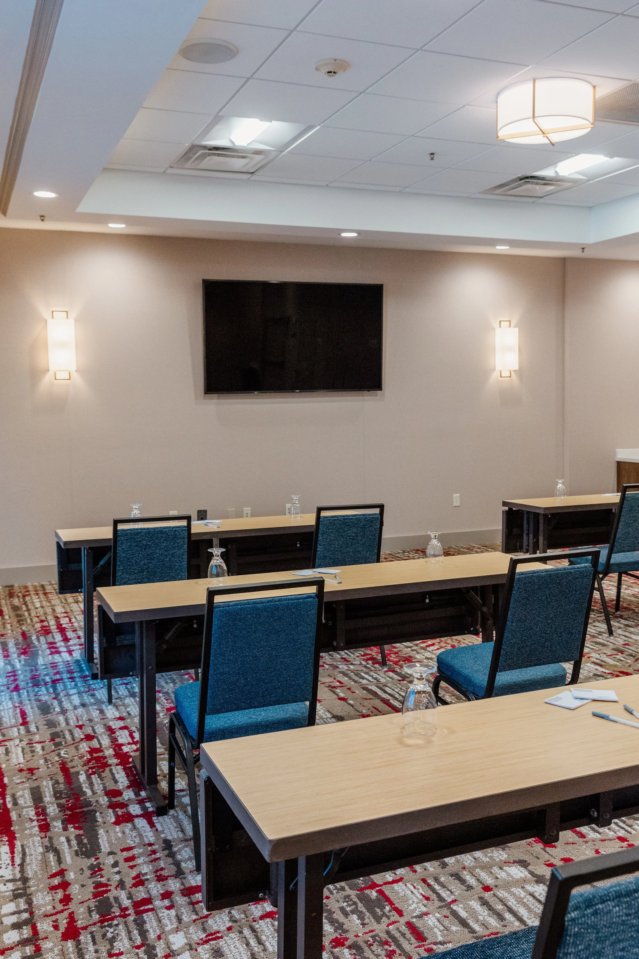 Empty conference room with tables and chairs, a large flat-screen TV on the wall, glasses on tables, and soft lighting.
