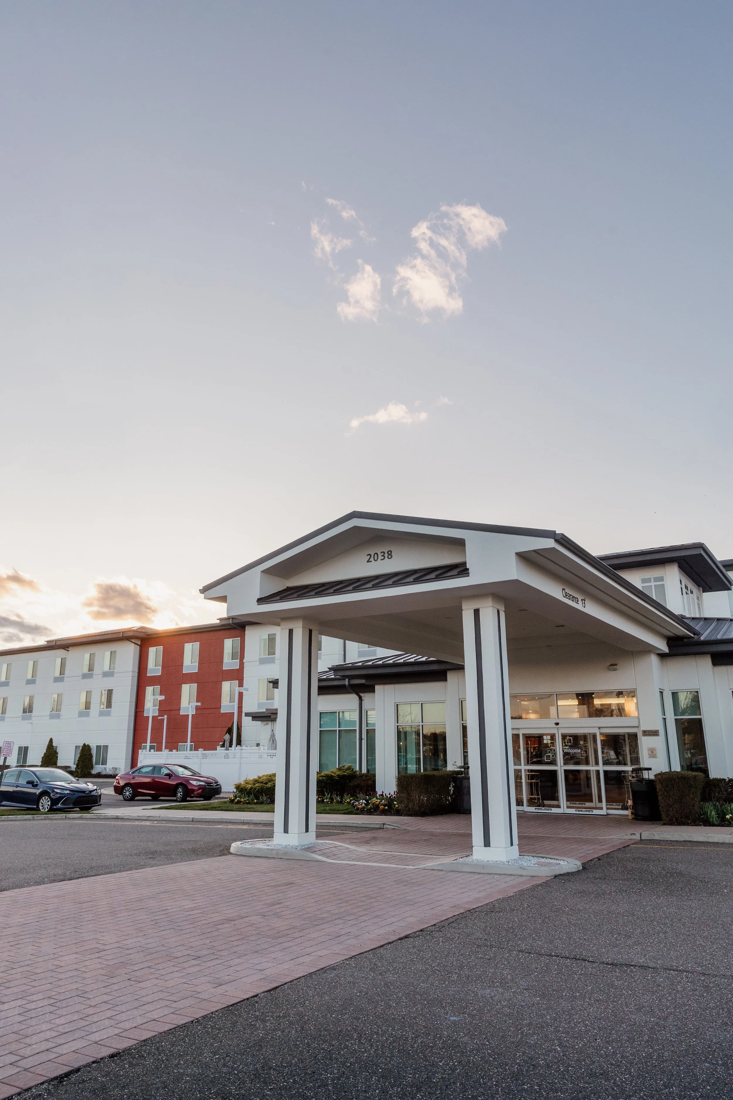 Exterior view of a modern hotel building during sunset, showing a covered entrance with glass doors, cars parked in front, and a clear sky with a few clouds.