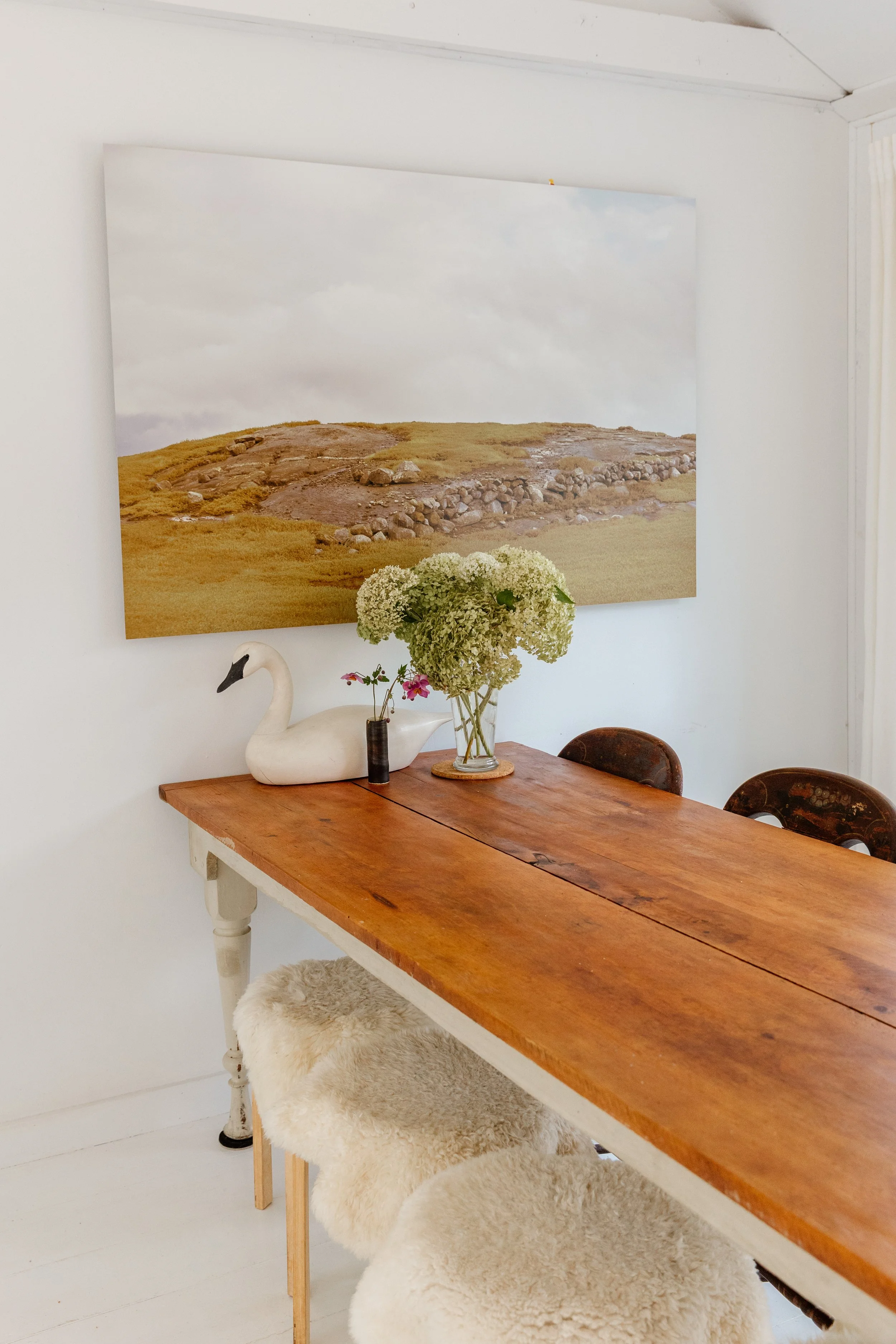 A wooden dining table with a large bouquet of green flowers in a glass vase, a white swan-shaped decorative object, and a small black vase with pink flowers. Behind the table, two wooden chairs and a sheepskin-covered stool are visible. A large lands