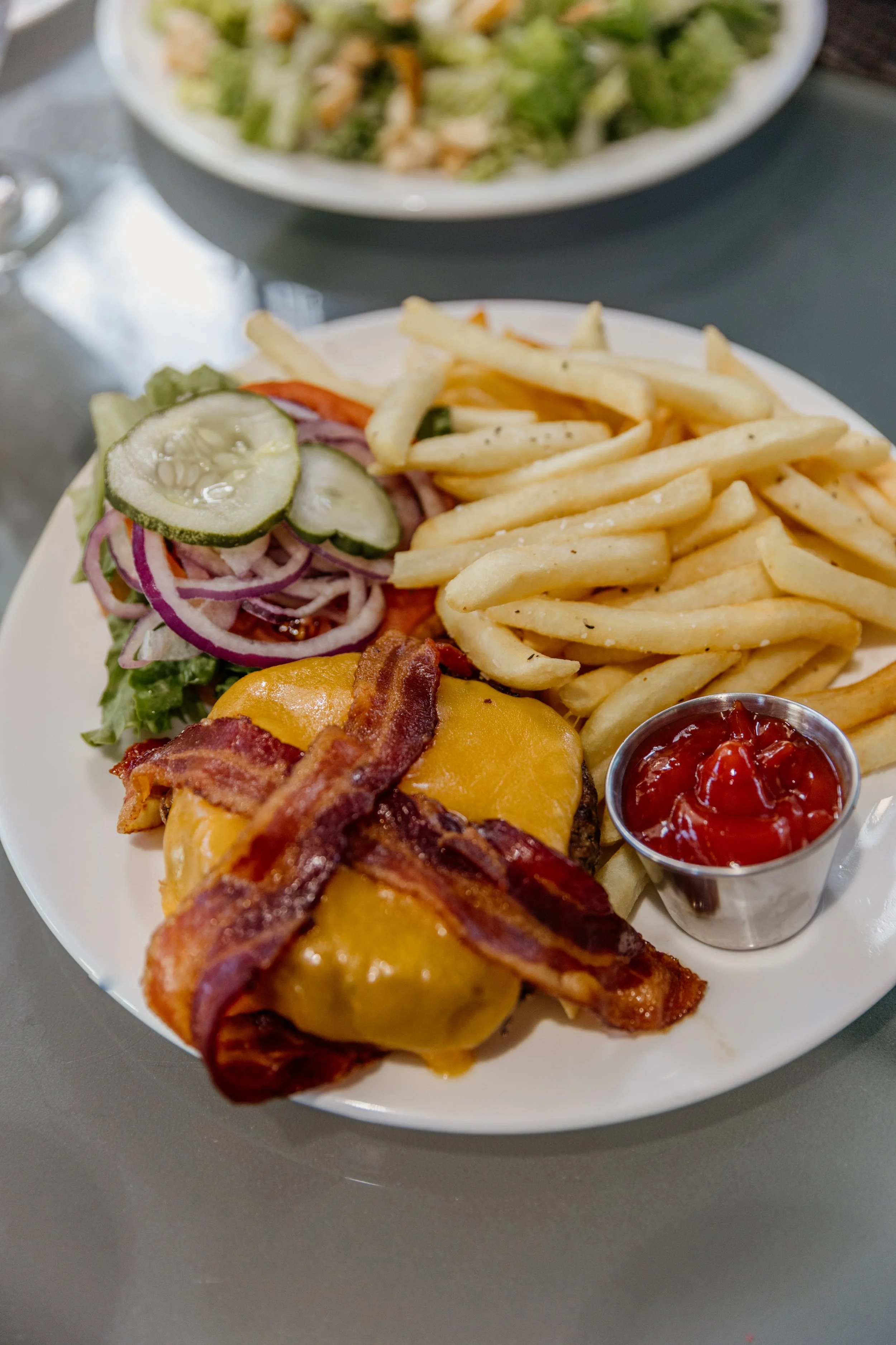 Plate with bacon cheeseburger, French fries, fresh salad, and a small cup of ketchup.