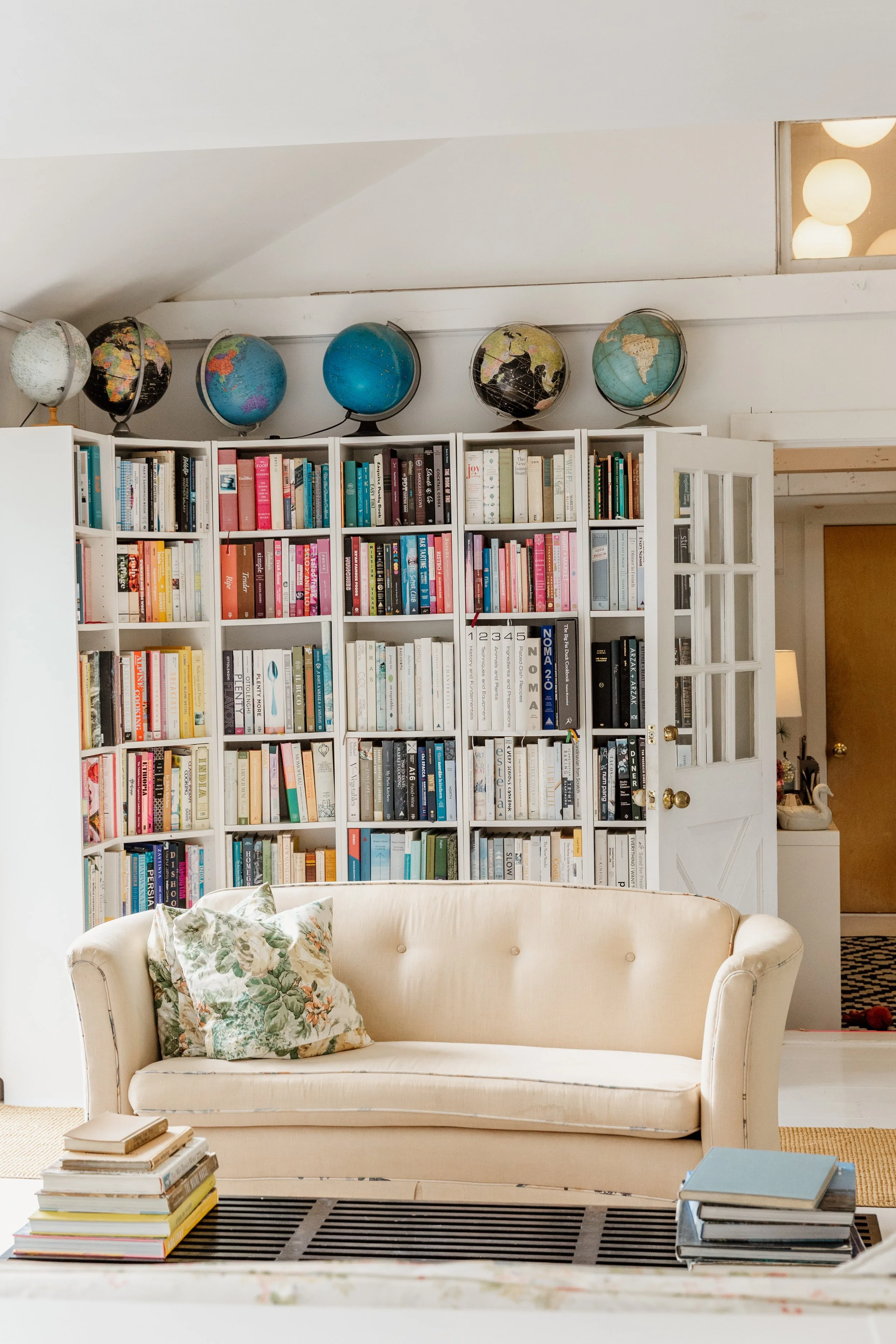 Living room with a cream-colored sofa, patterned cushions, a coffee table with stacked books, and a white bookshelf filled with books, with globes on top of the shelf.