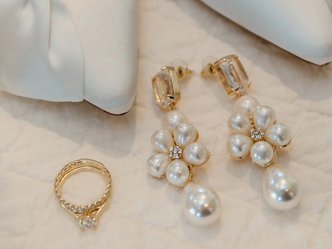 Pair of white pearl drop earrings with gold accents, a gold ring with small diamonds, on a white fabric surface, with a light-colored purse or cloth in the background.