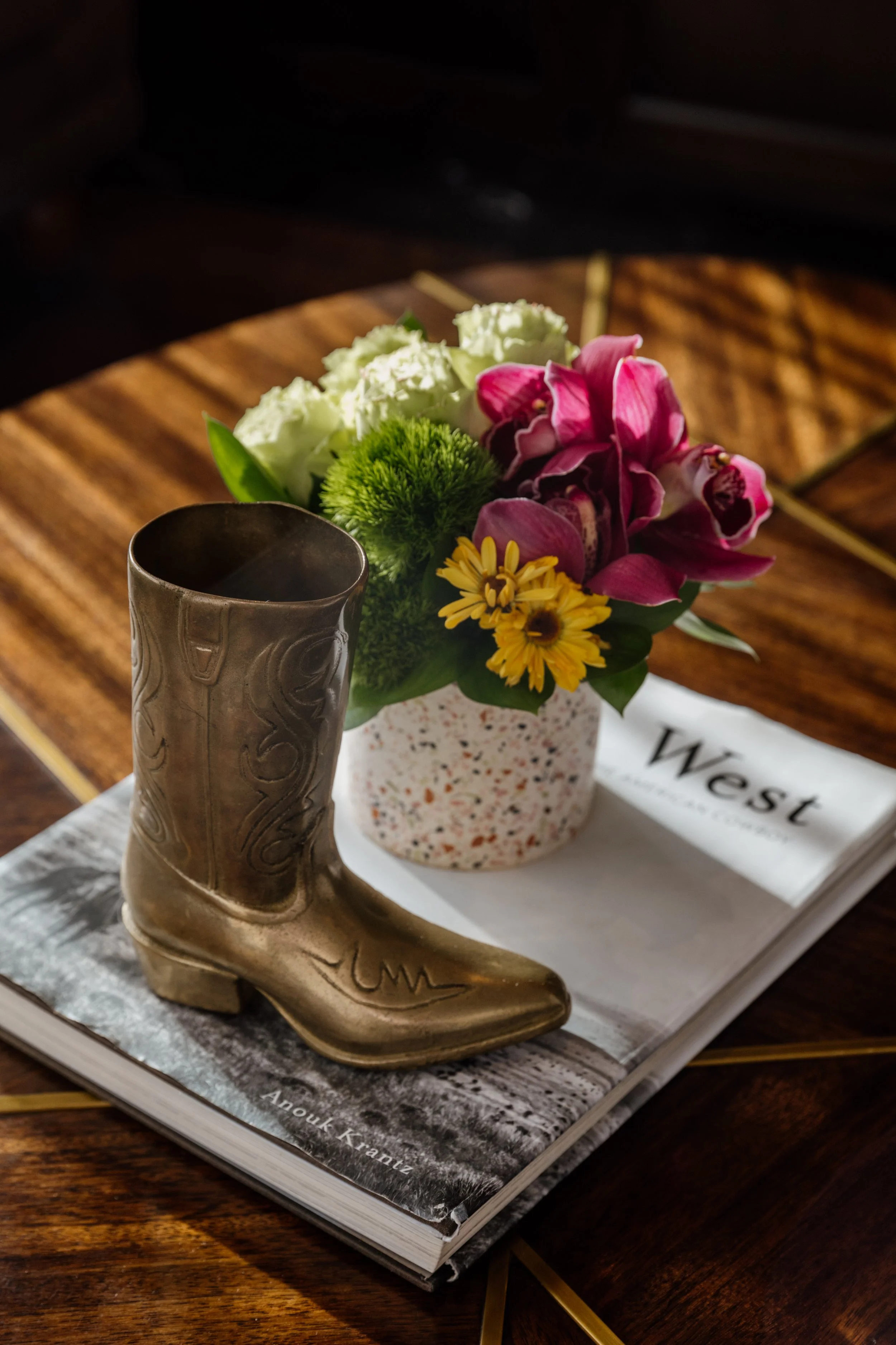A small decorative boot-shaped container placed on a magazine, next to a flower arrangement in a speckled vase on a wooden table.