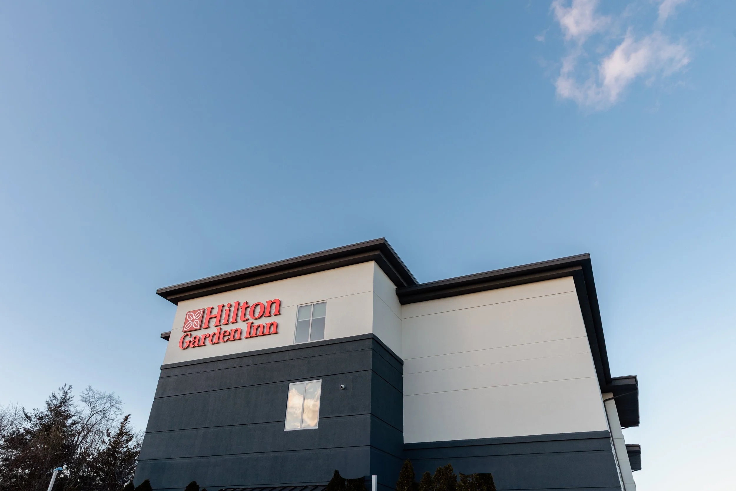 Exterior view of a Hilton Garden Inn hotel building with a red Hilton Garden Inn sign on the upper left corner, against a clear blue sky with a few clouds.