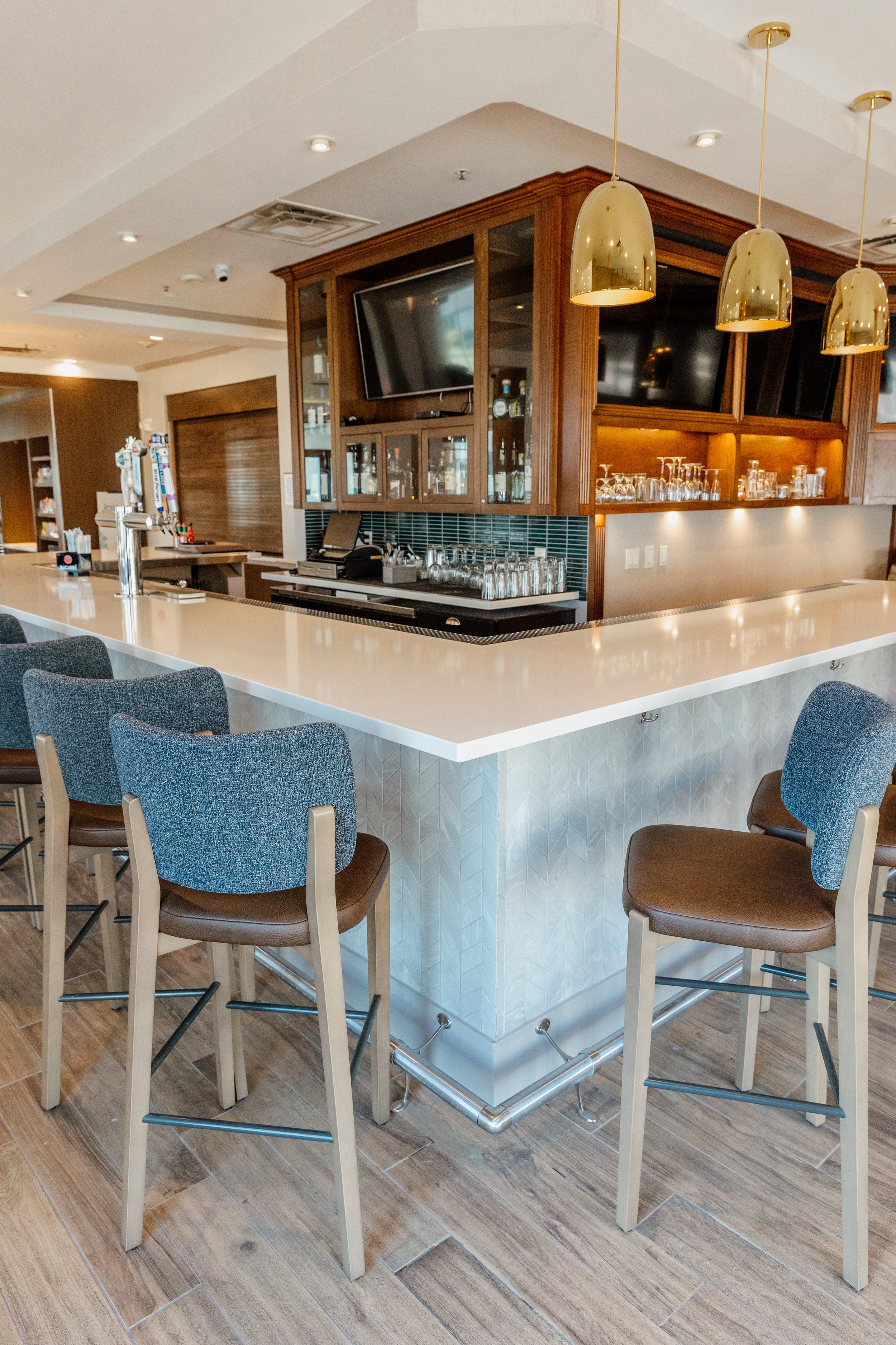 A bar with a white countertop, brown bar stools with blue upholstery, wooden shelving with glasses, gold pendant lights, and television screens.