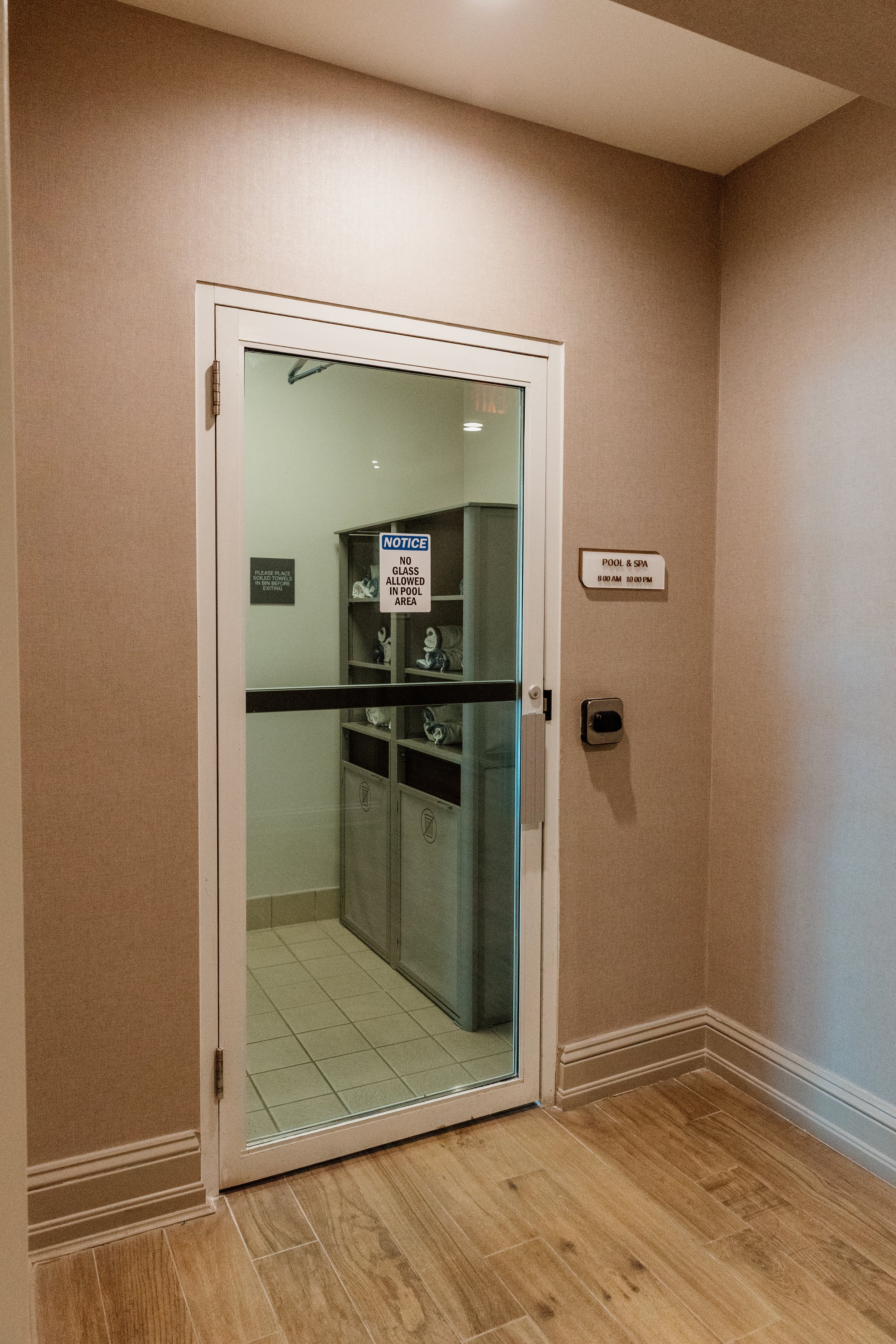 A glass door labeled Pool & Spa leads to a room with shelves holding towels and lockers, with signs indicating no glass allowed in the pool area and the pool and spa hours.