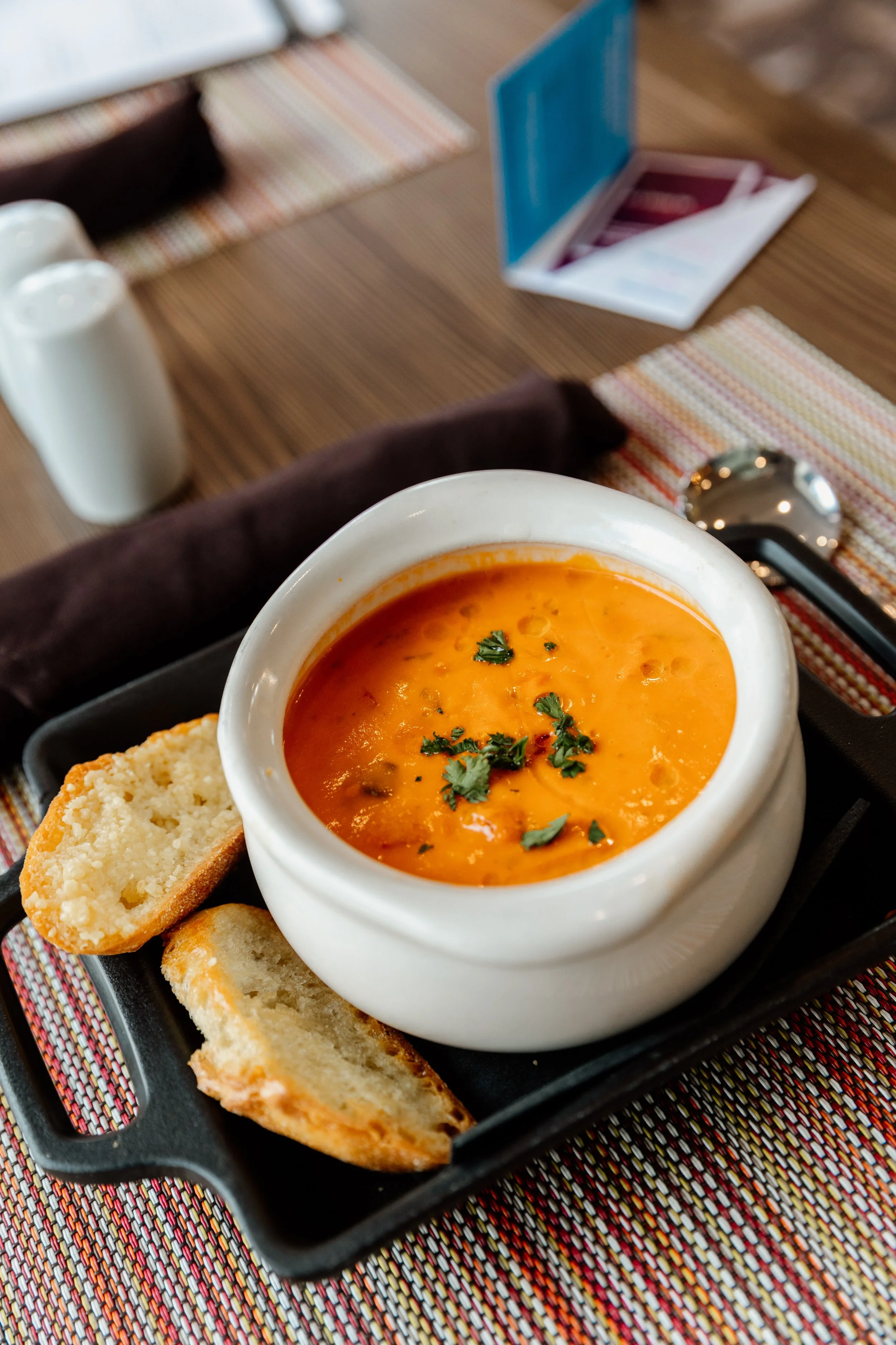 A bowl of orange-colored soup garnished with herbs, served with sliced bread on a tray at a table.