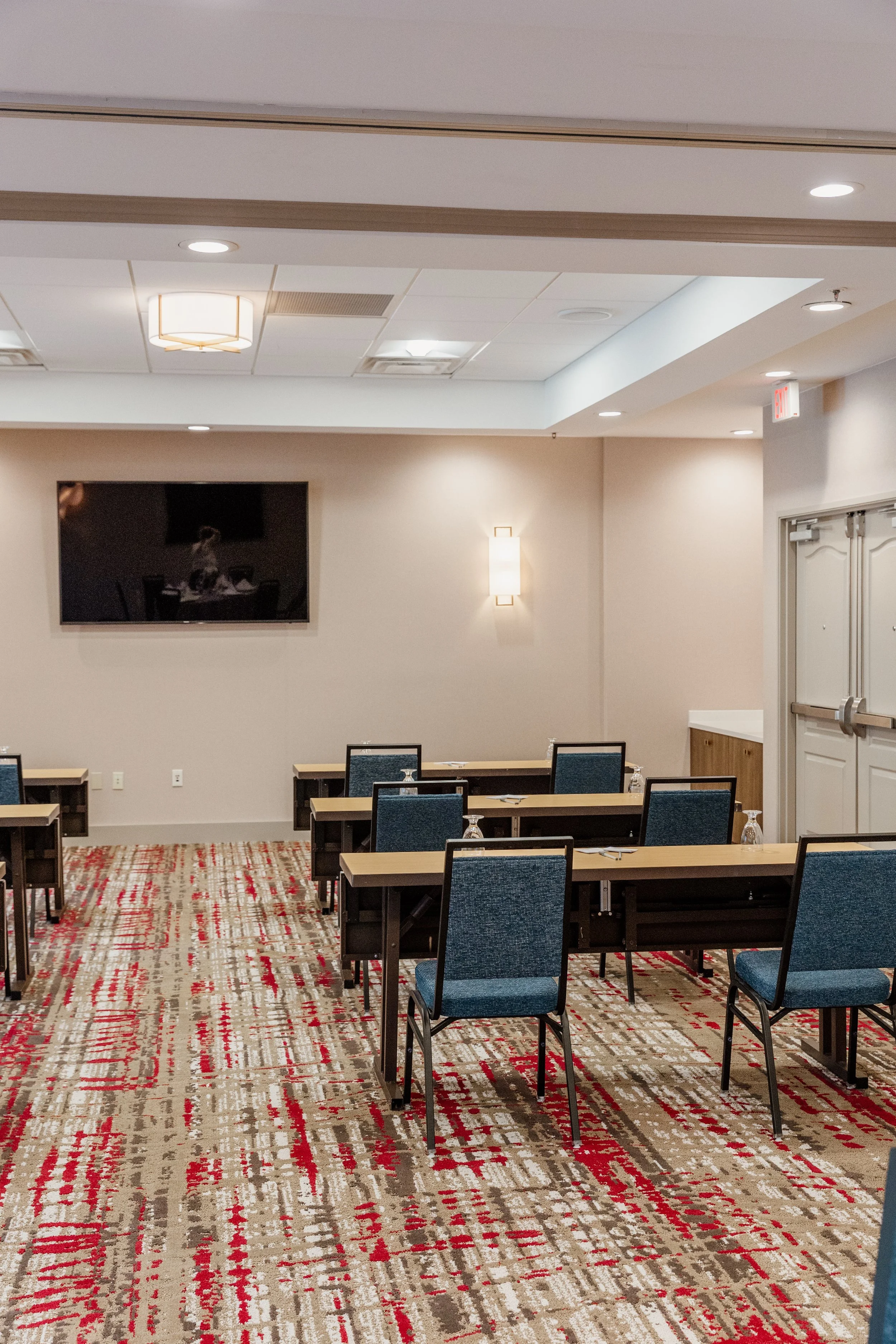 Empty conference room with tables, chairs, a wall-mounted TV, and ceiling lights.