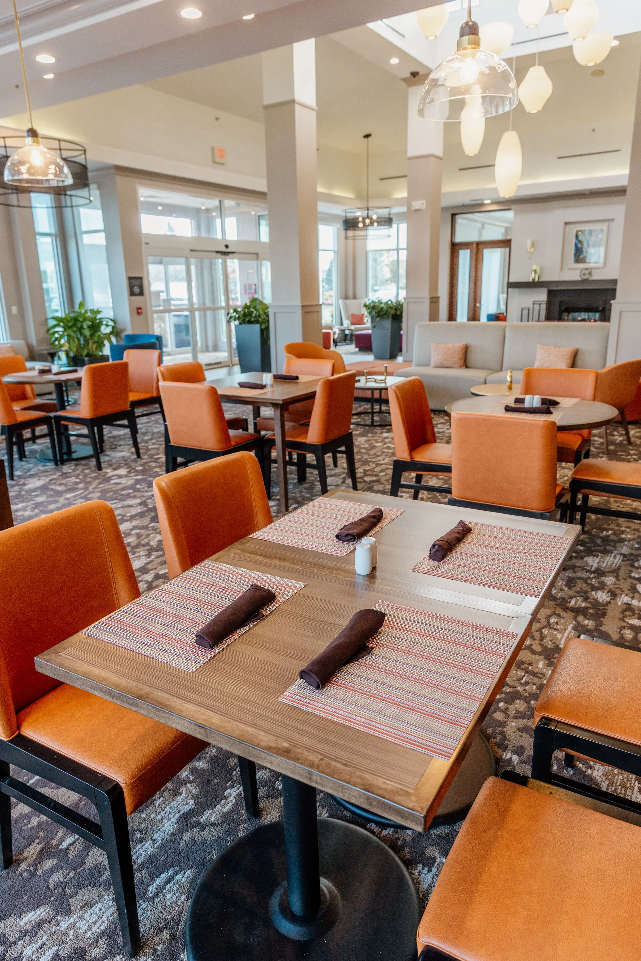 Empty restaurant with wooden tables, orange chairs, patterned carpet, and large windows letting in natural light.