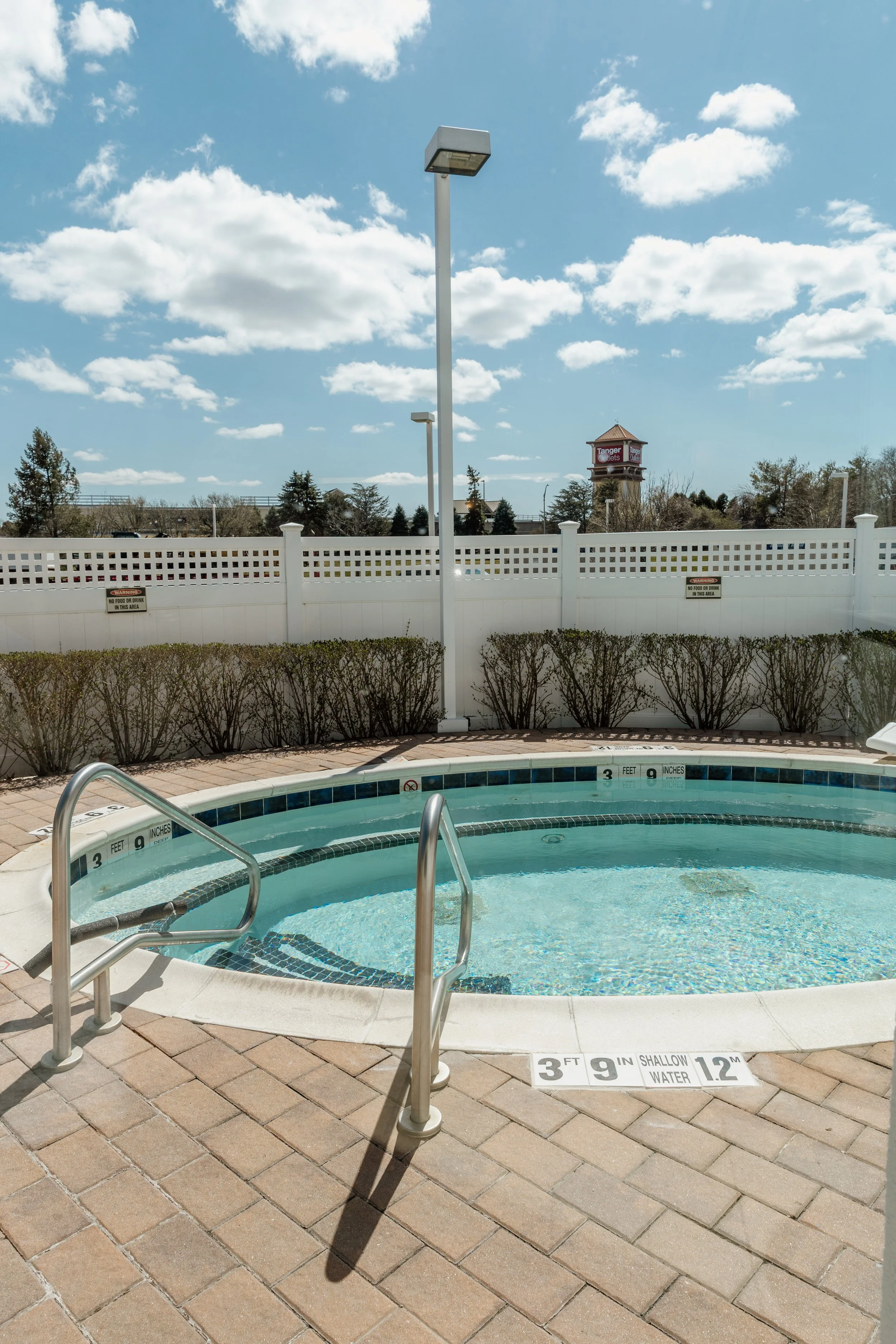 An empty outdoor swimming pool with clear water, metal handrails, and a brick-paved deck, enclosed by a white fence, under a partly cloudy blue sky.