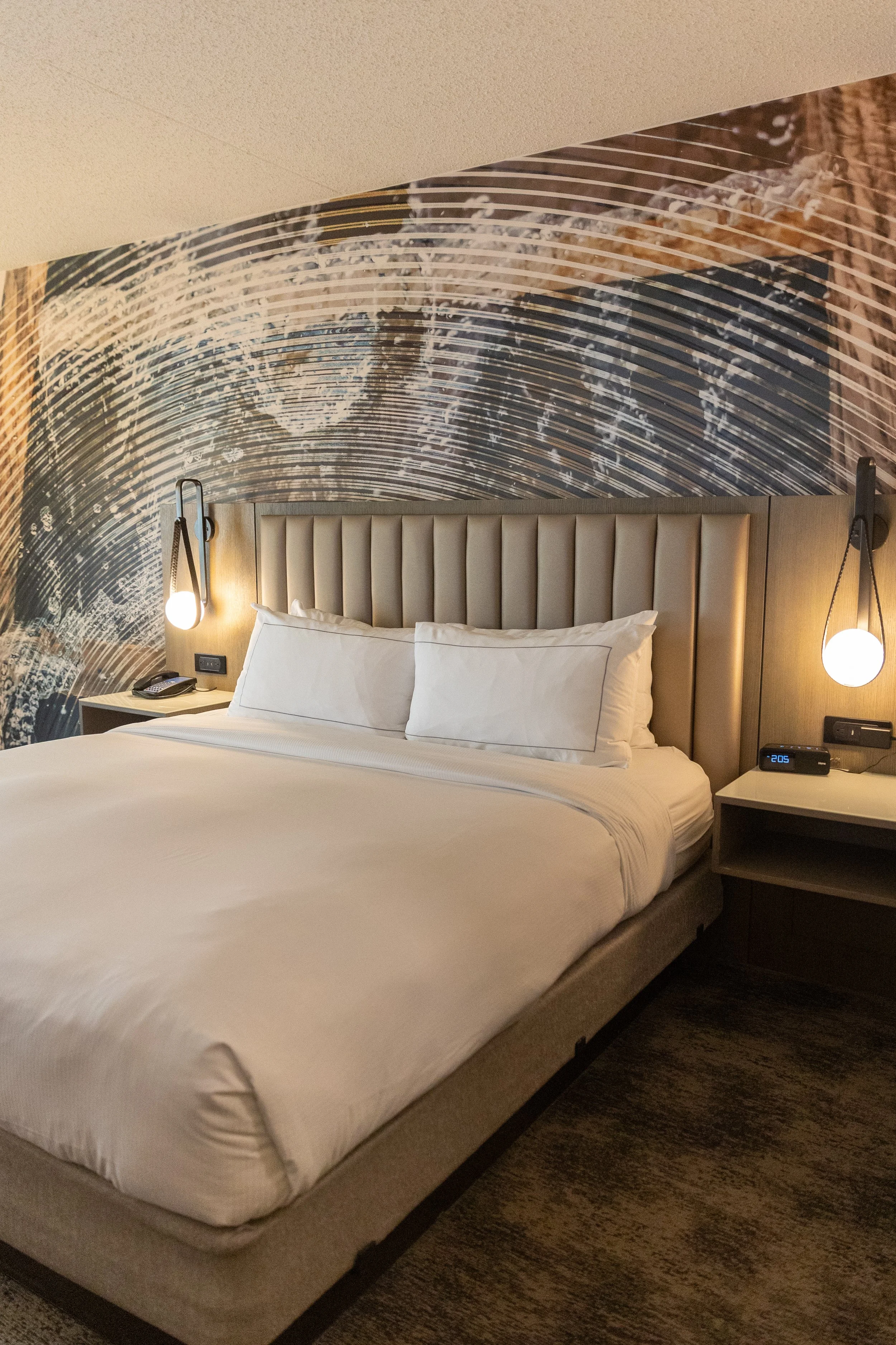 Hotel room with a large bed, beige upholstered headboard, white bedding, two pillows, wall-mounted lamps, digital alarm clock, telephone, and an artistic mural wall behind the bed.