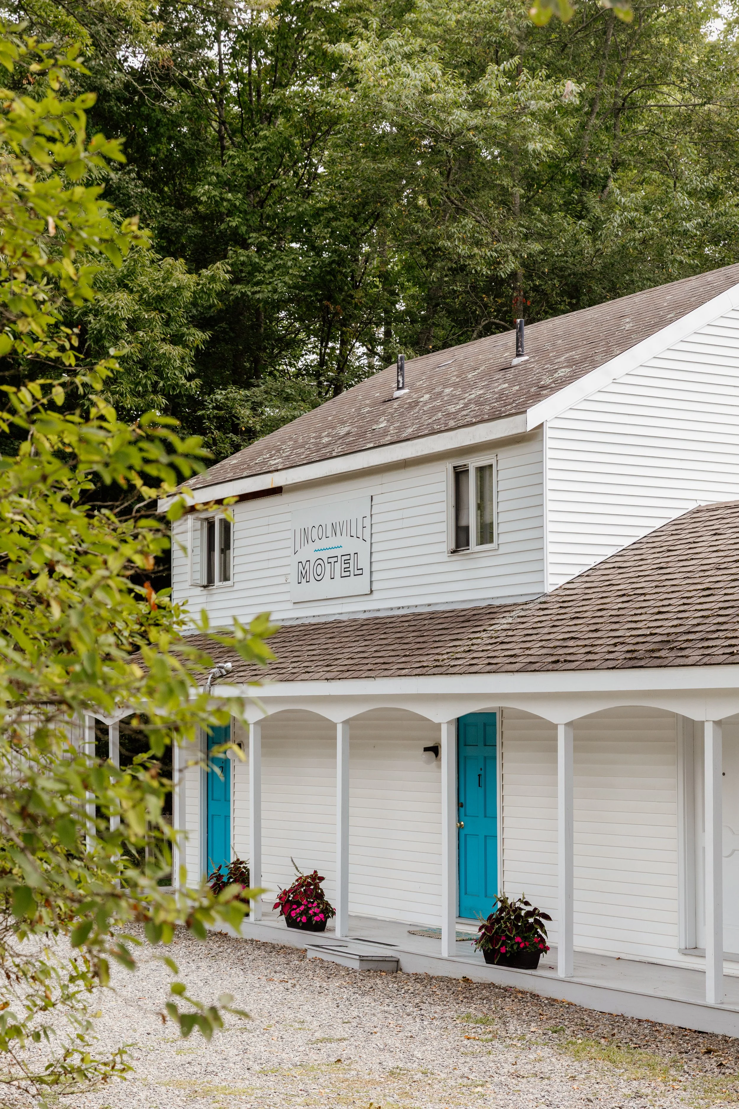 White building with a sign that reads 'Lincolnville Motel,' featuring a front porch with blue doors and nearby flower pots with pink flowers, surrounded by green trees.