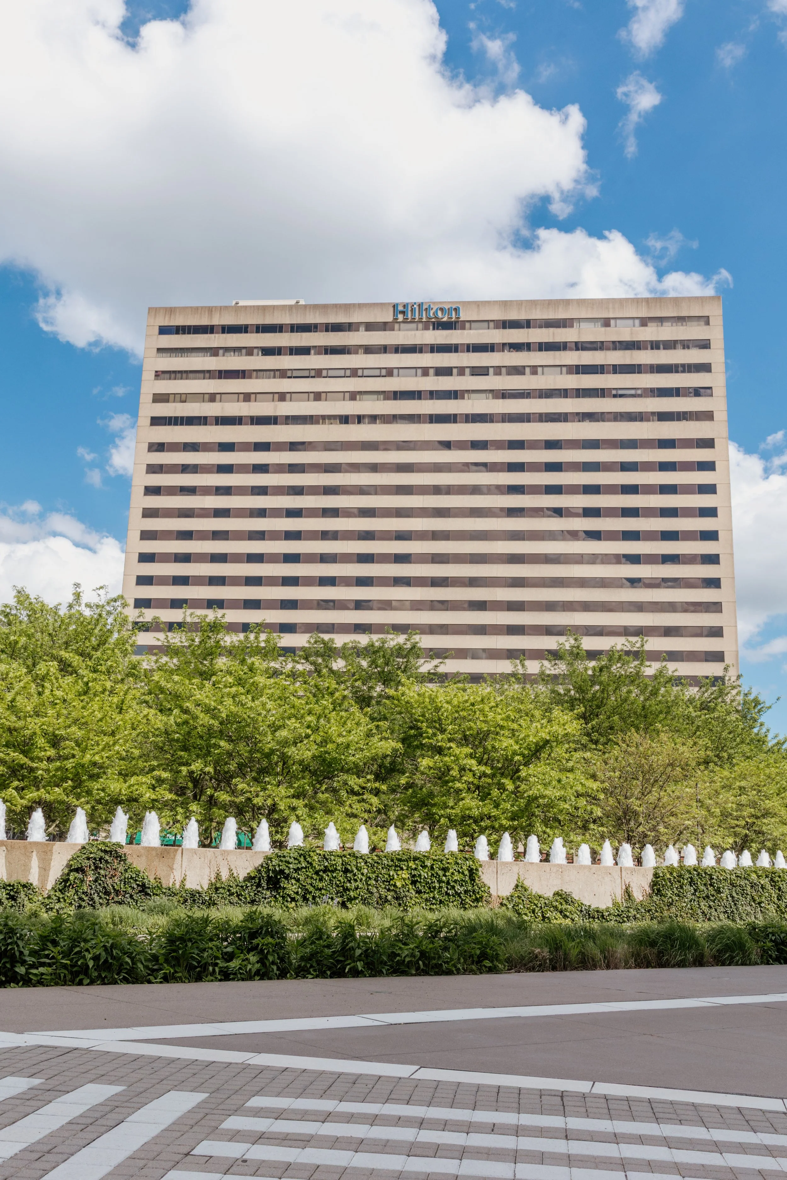 A tall Hilton hotel building with a blue sky and white clouds above and green trees and bushes with a fountain in front.