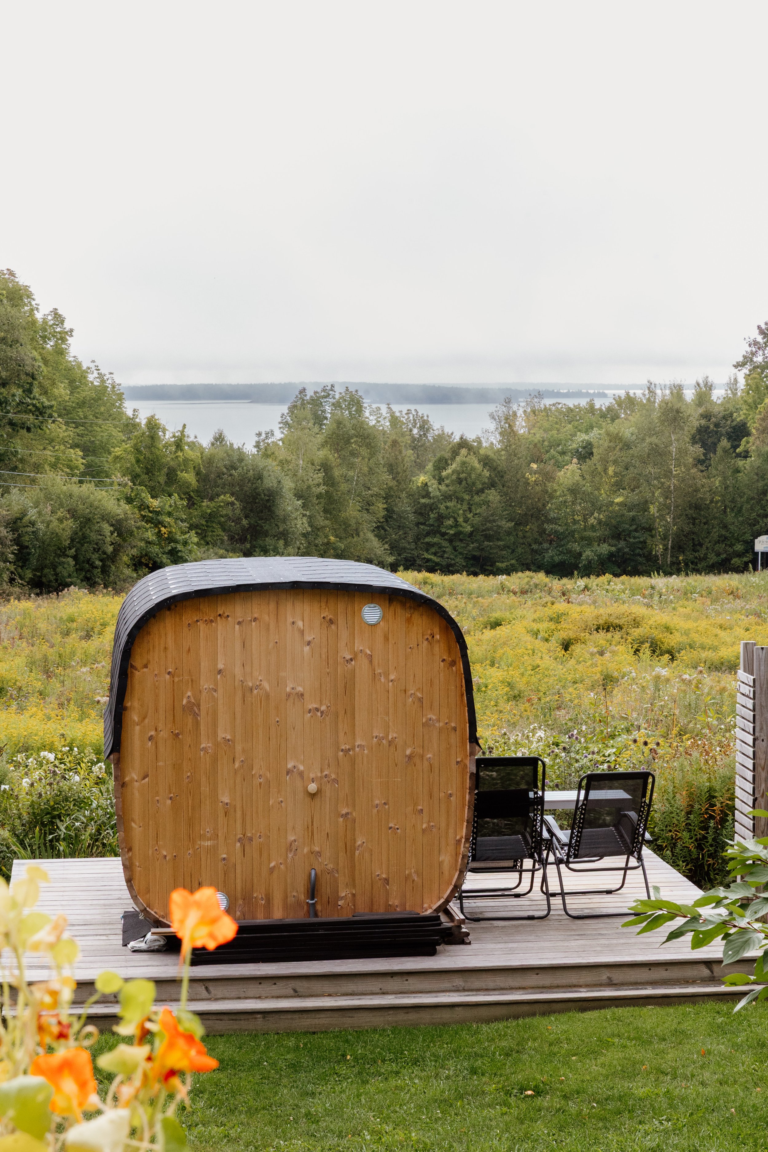 A backyard with a wooden hot tub on a deck, overlooking a field with flowers, trees, and a body of water in the distance under an overcast sky.